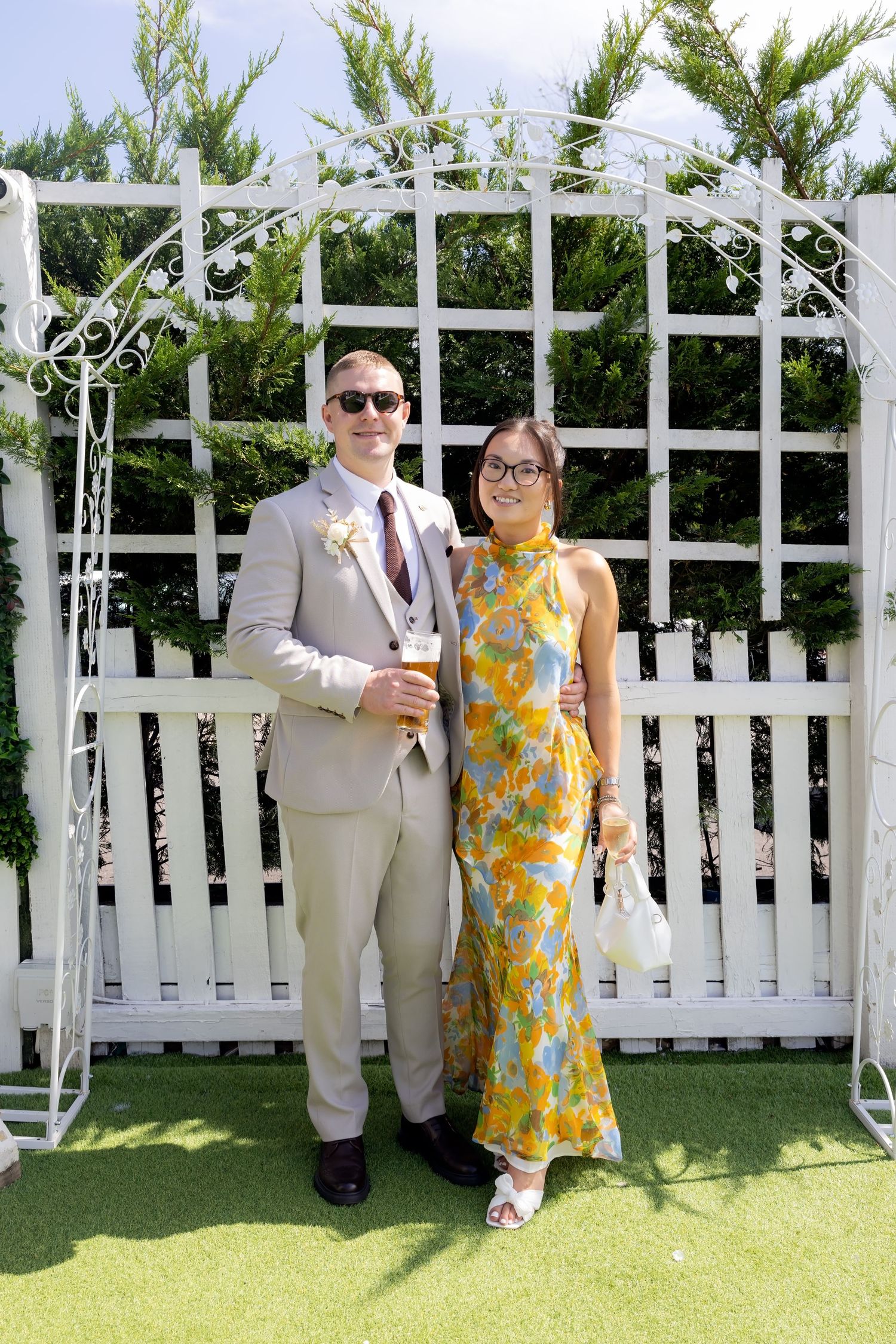 Wedding guest couple posing before white garden trellis, man in grey suit, woman in colorful floral dress.