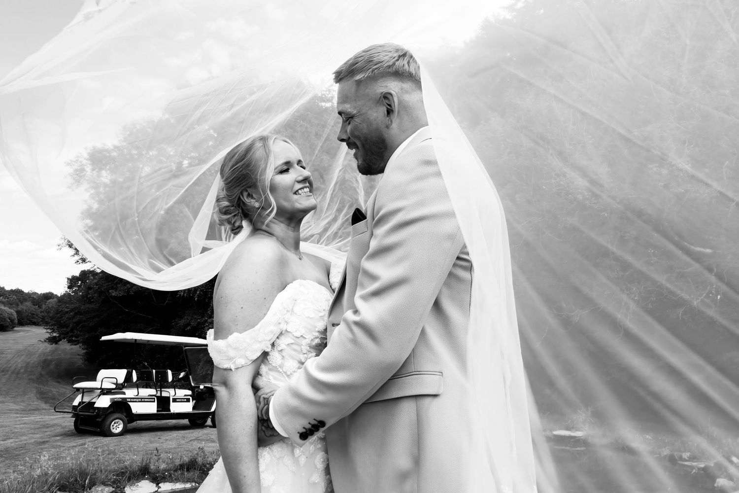 Black and white photo of bride and groom smiling under flowing veil with golf cart behind them.