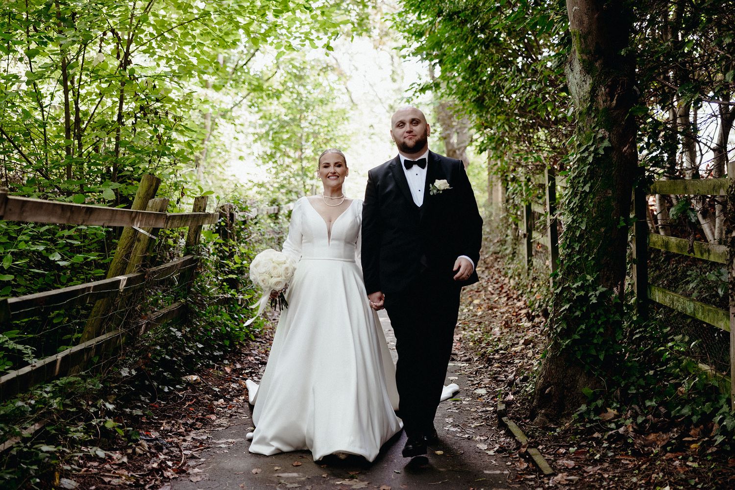 Bride in white gown and groom in black tuxedo walking hand in hand through woodland path.
