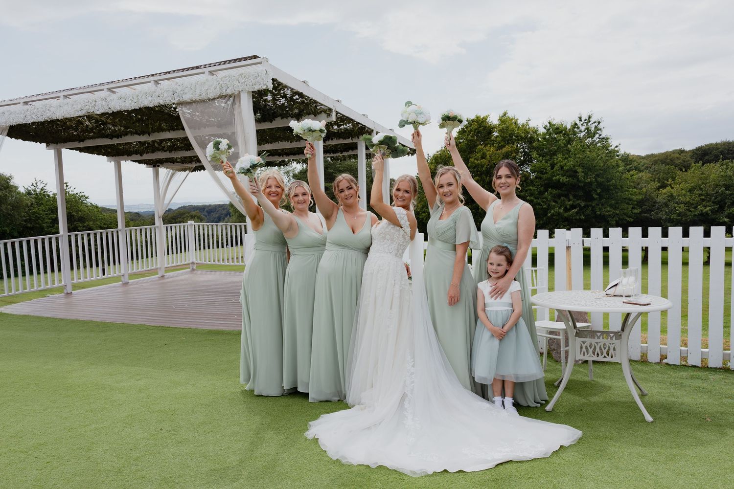 Bride and five bridesmaids in sage green dresses raising glasses joyfully on a sunny wedding day.