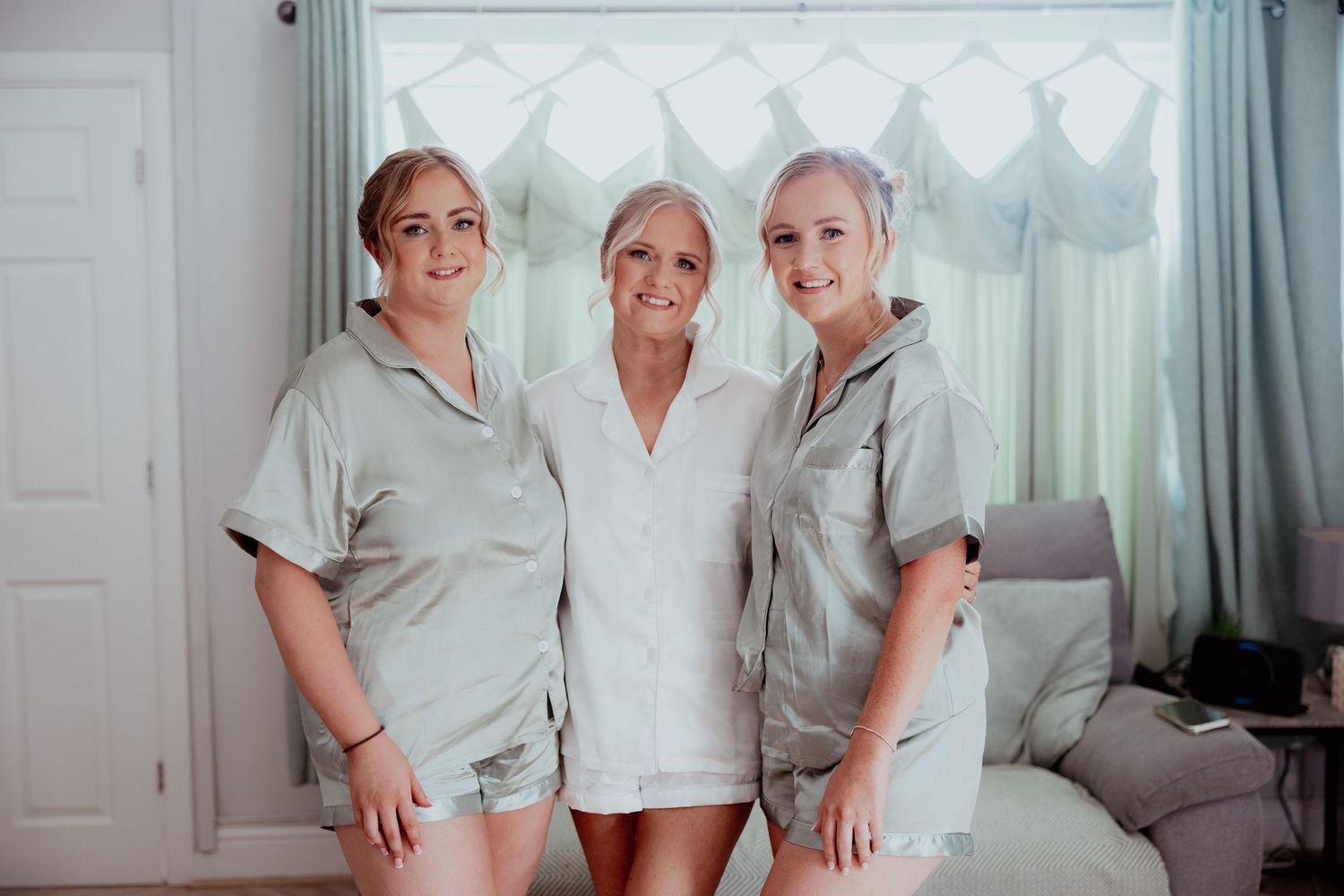 Bride and two bridesmaids smiling together in matching satin pyjamas during wedding morning preparations.