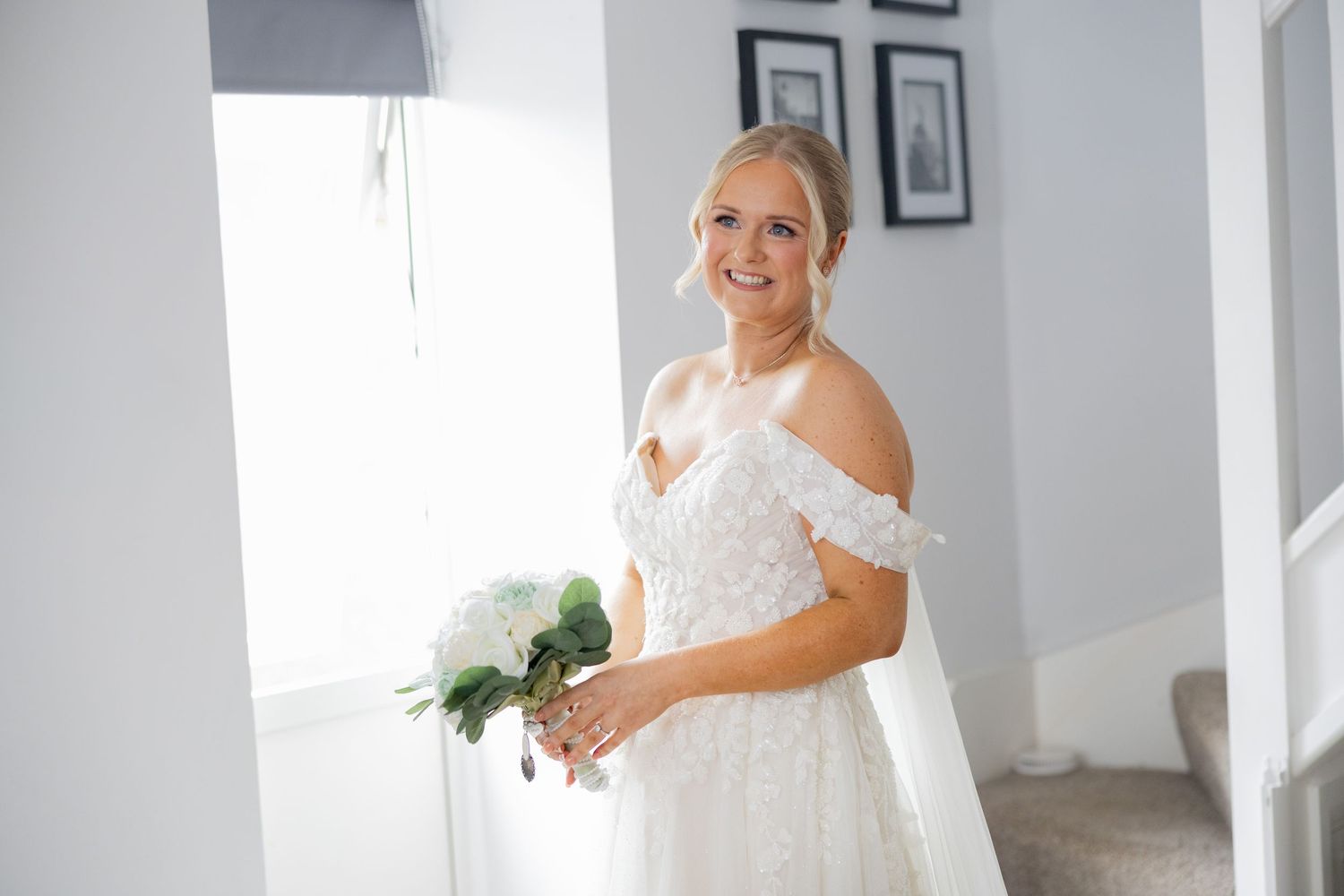 Radiant blonde bride in a floral lace off-shoulder wedding gown holding a white bouquet indoors.