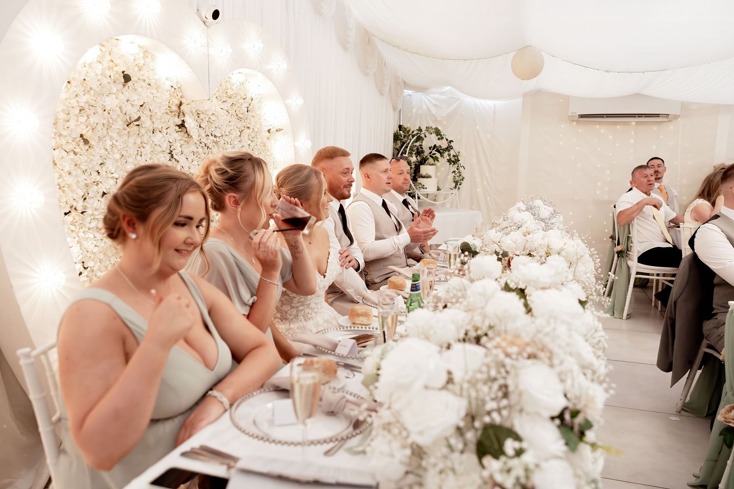 Wedding party enjoying reception dinner at elegantly decorated table with white floral arrangements.