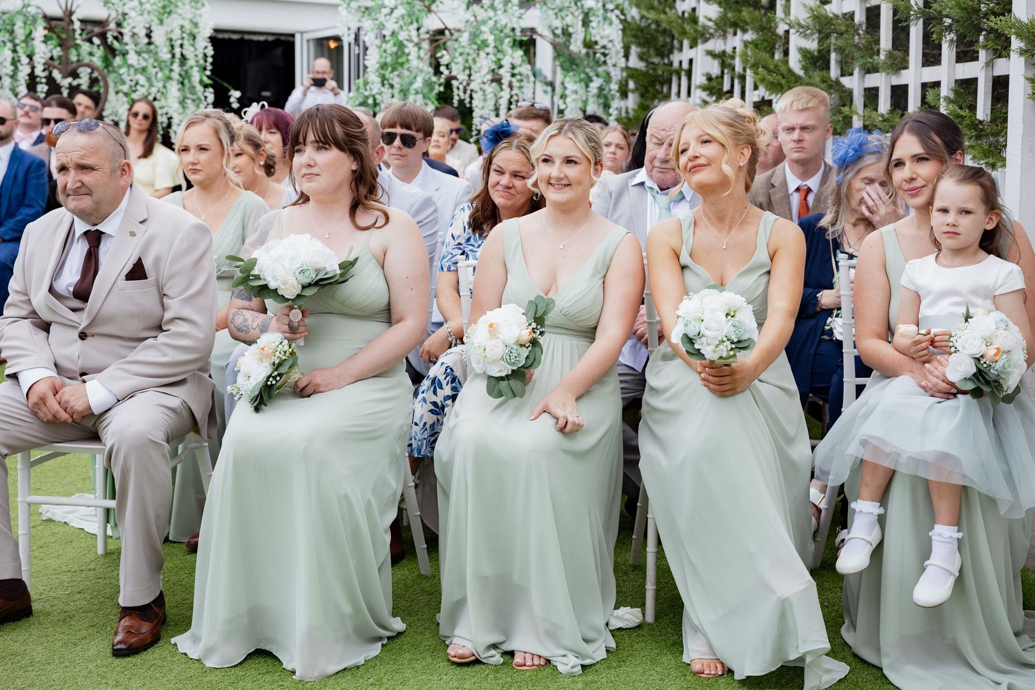 Bridesmaids in sage green dresses holding white bouquets seated at an outdoor wedding ceremony. south Wales weddings photography.