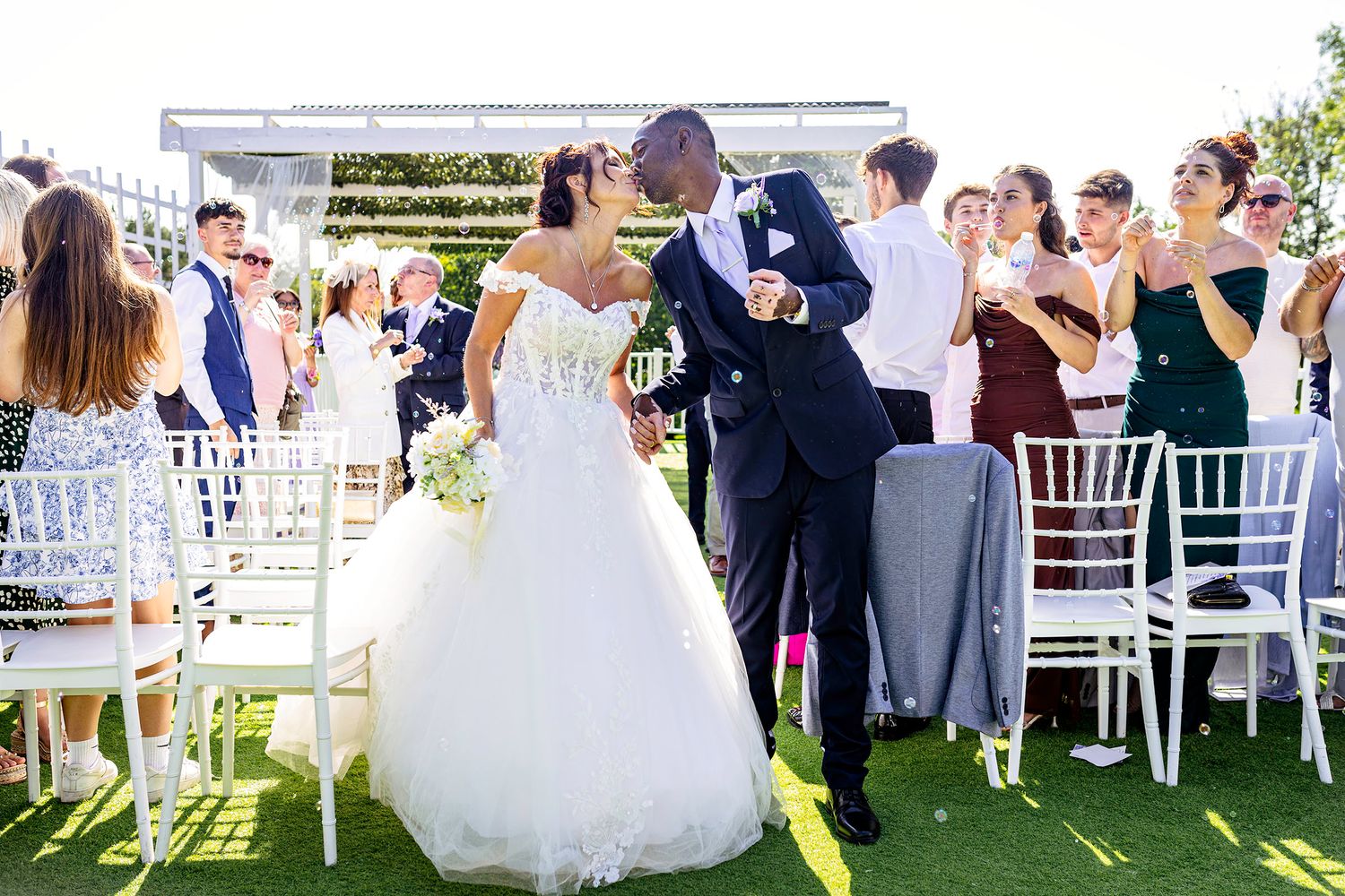 A bride and groom share a kiss at an outdoor wedding ceremony surrounded by guests on a sunny day. South Wales wedding photography