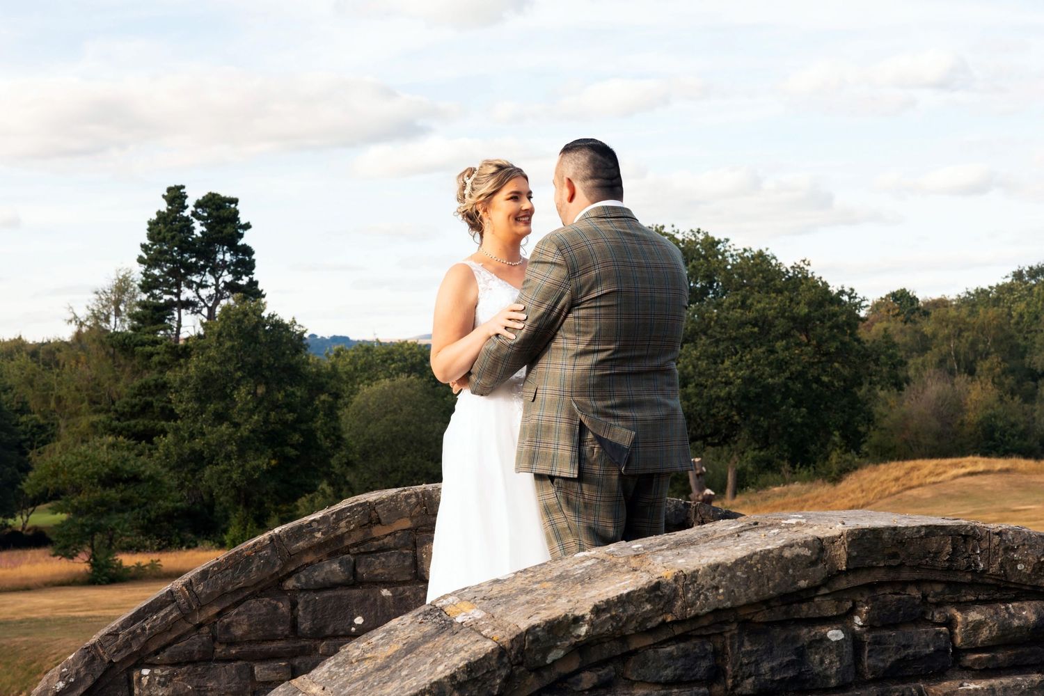 Bride and groom share a joyful moment on stone ruins with scenic greenery and cloudy sky backdrop at outdoor wedding.