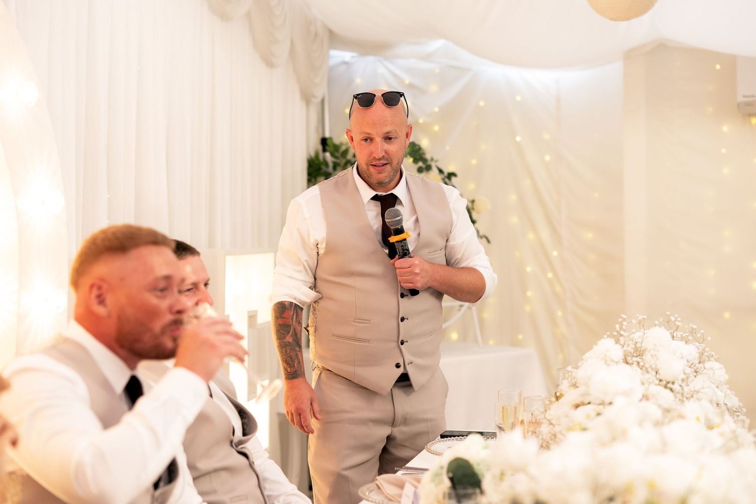 Man in grey waistcoat delivers wedding speech holding microphone at elegant reception.