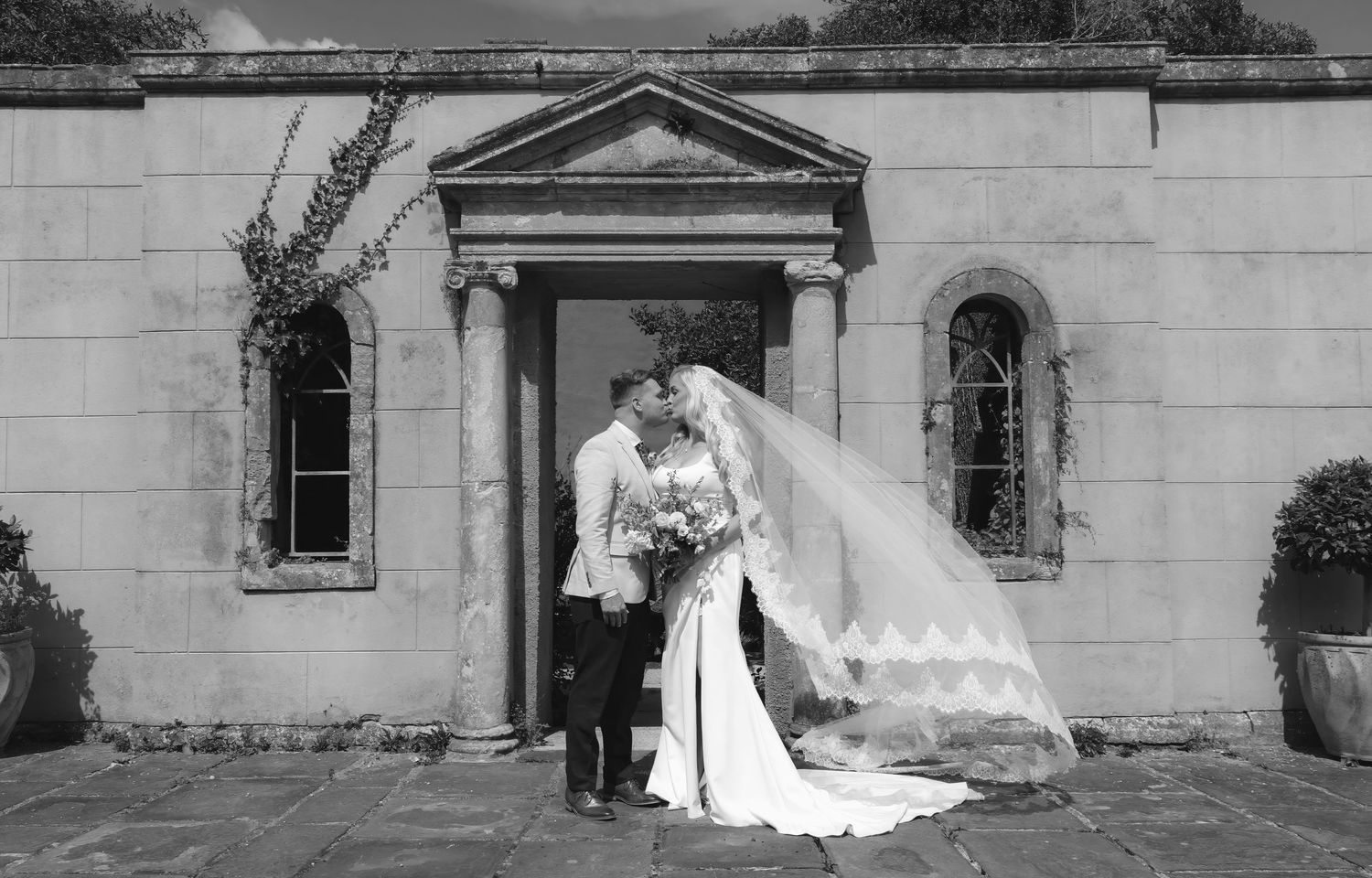 Black and white wedding photo of bride and groom kissing outside elegant stone building with flowing veil.