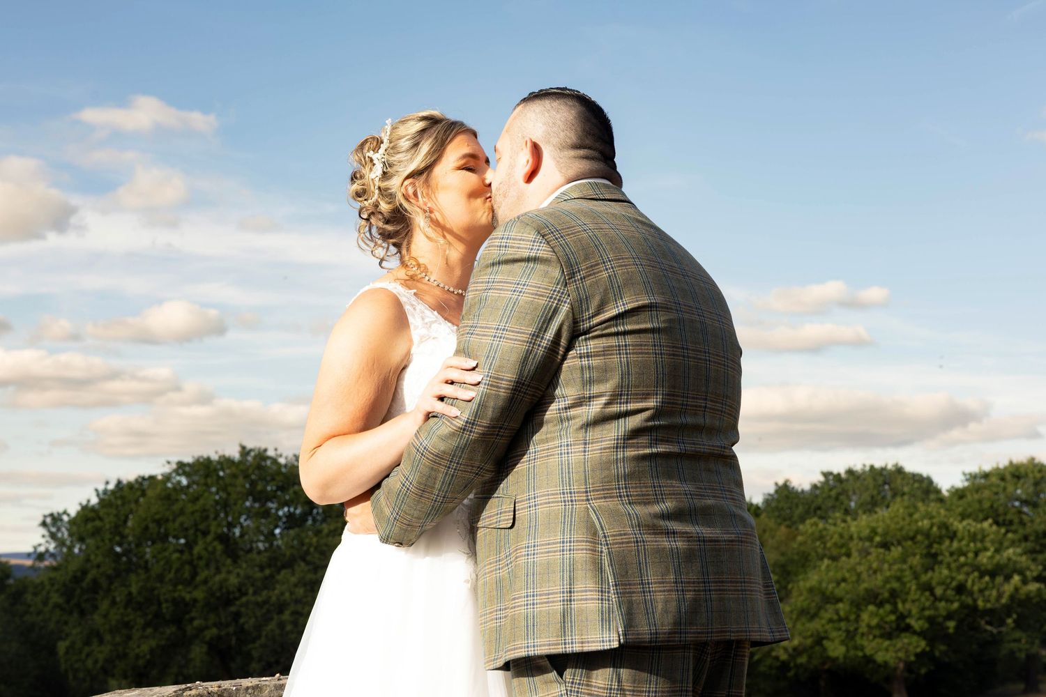 Bride and groom share a romantic kiss outdoors with blue sky and green trees behind them on their wedding day.