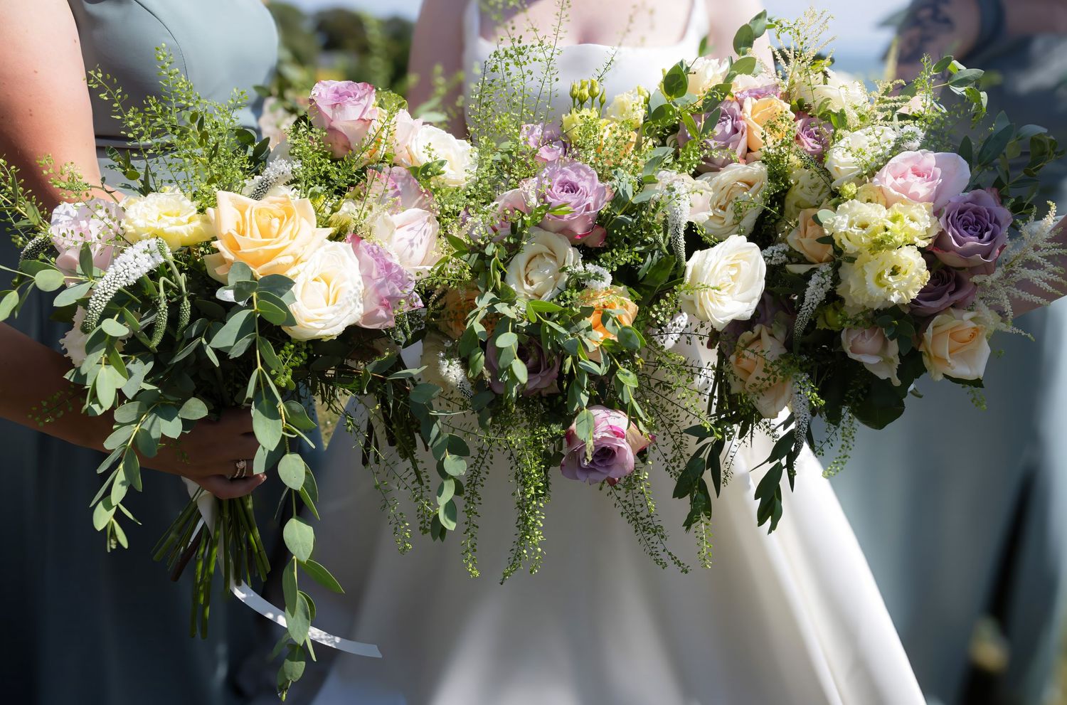 Bridesmaids holding lush cascading bouquets of pastel roses, ranunculus, and greenery at a wedding.