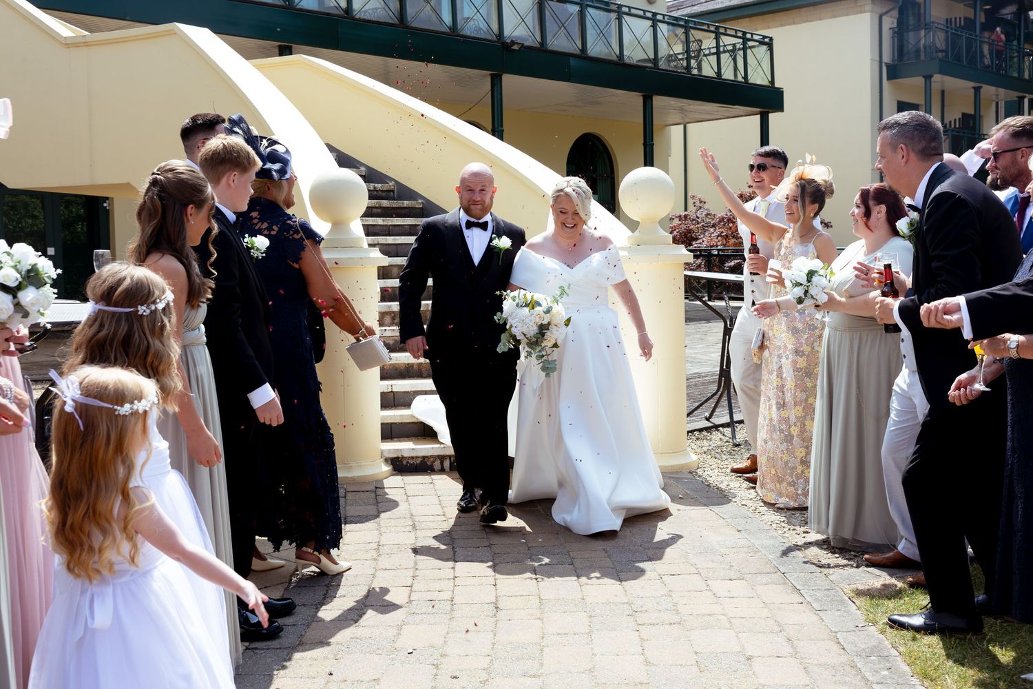 Bride and groom walk through cheering guests outdoors at a sunny wedding ceremony venue.