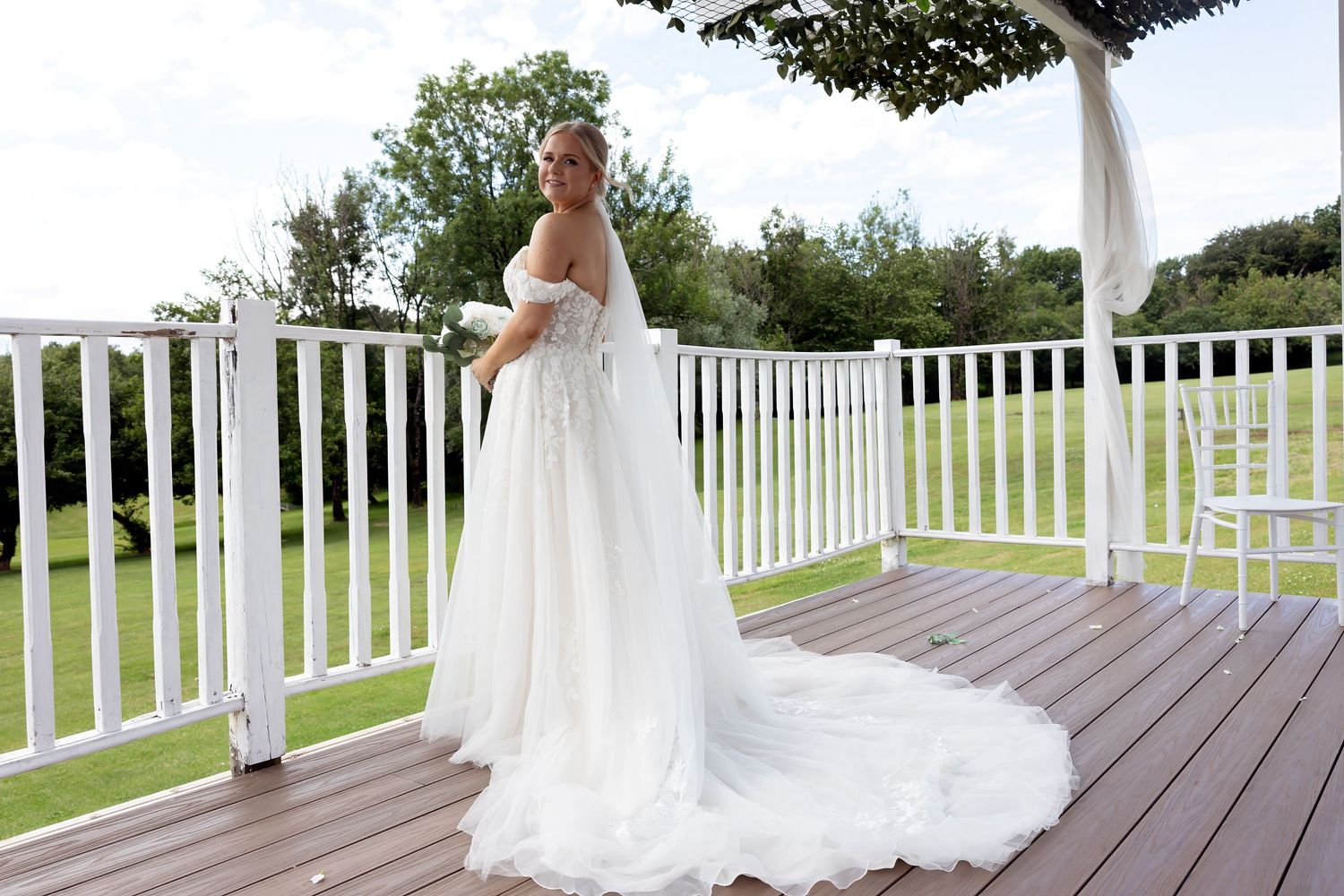 Bride in flowing white off-shoulder wedding dress with long train standing on wooden deck.