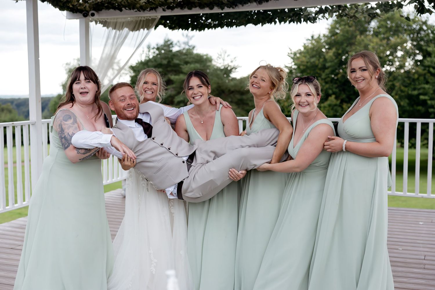 Bridesmaids in sage green dresses playfully lifting groom while bride laughs at outdoor wedding reception.