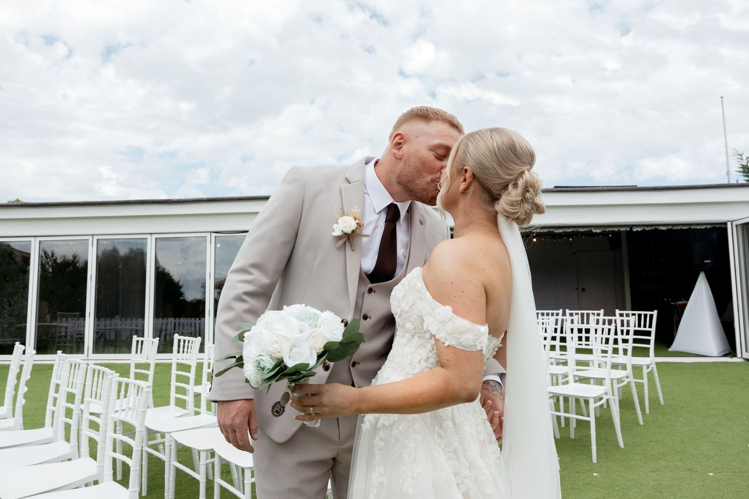 Bride and groom share a kiss outdoors at a wedding venue with white chairs arranged on green grass. welsh wedding photography.