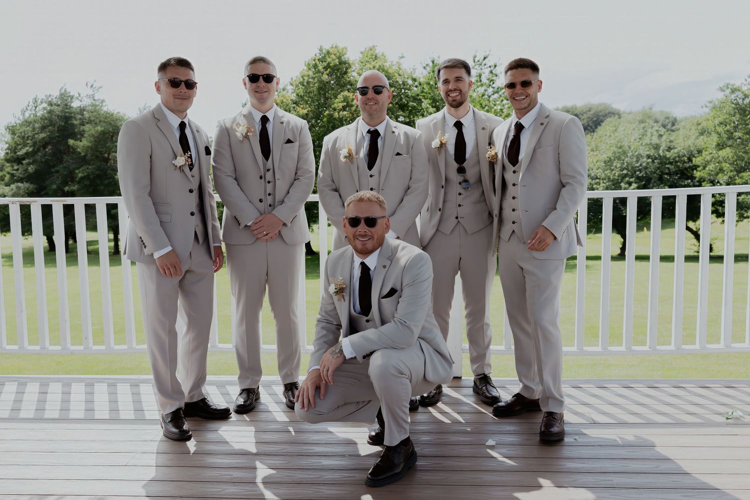 Groom and groomsmen in matching grey suits and sunglasses pose on outdoor deck at sunny wedding venue.