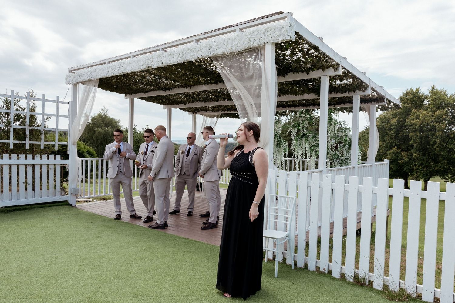 Groomsmen in grey suits waiting under white pergola at outdoor wedding ceremony.