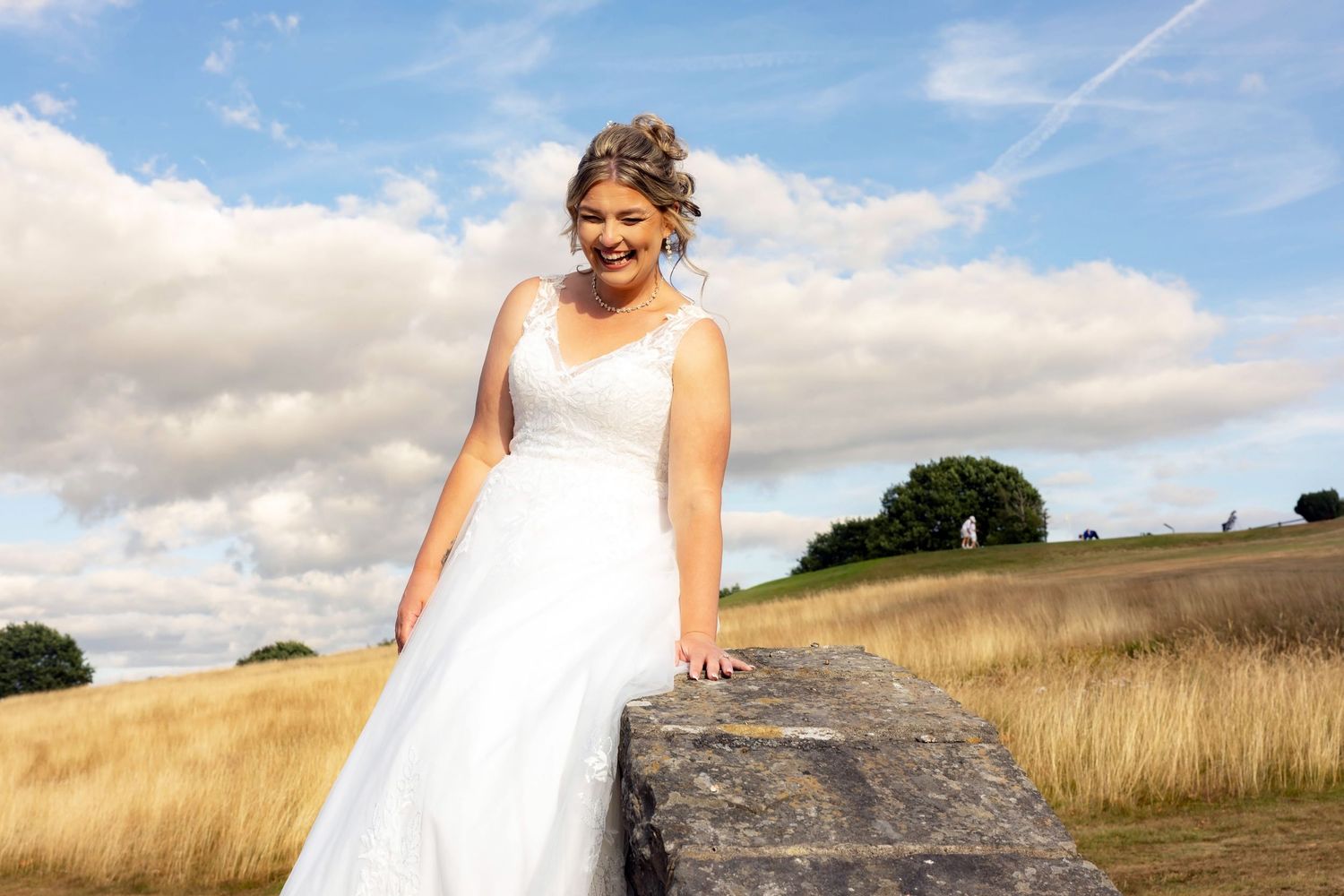 Bride in white lace gown posing on stone wall in sunny countryside landscape.