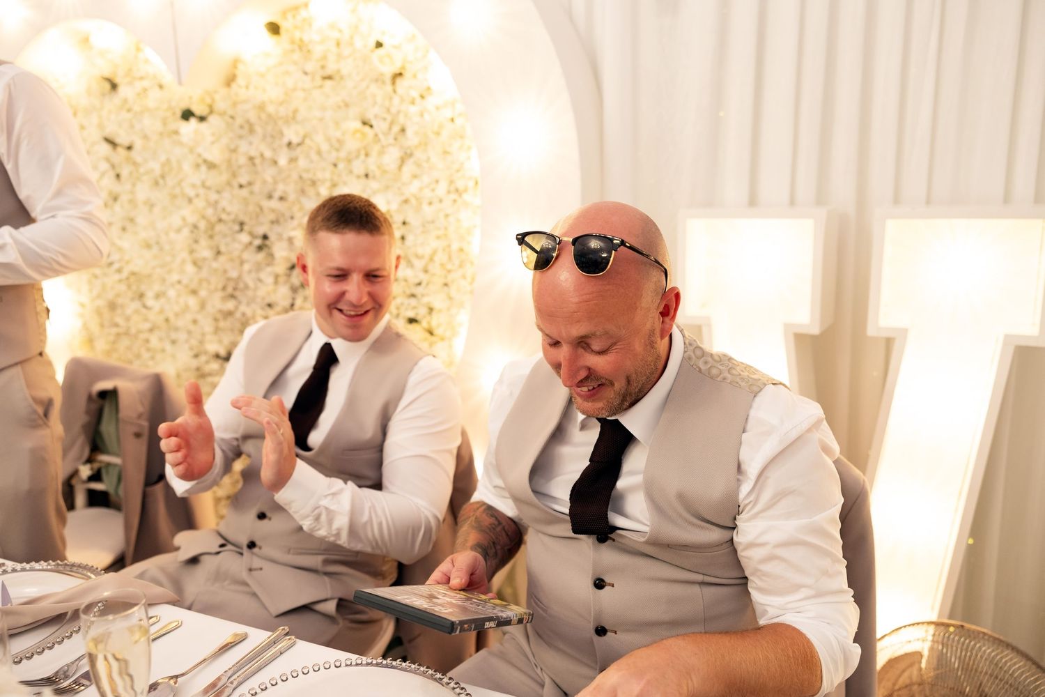 Two men in grey waistcoats laughing together at wedding reception head table.