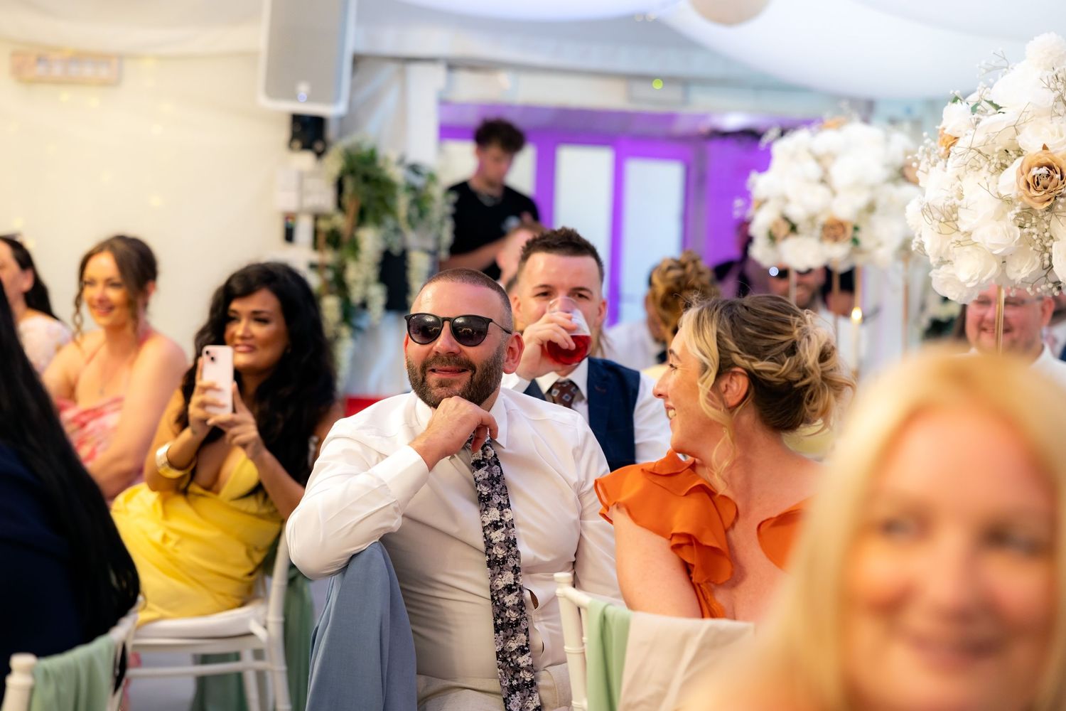 Wedding guests laughing and celebrating during speeches at a decorated reception venue.