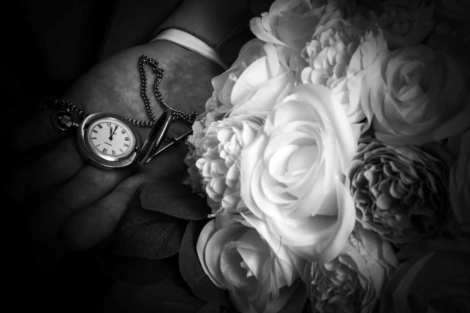 Black and white close-up of a groom holding a pocket watch beside a white bridal bouquet.