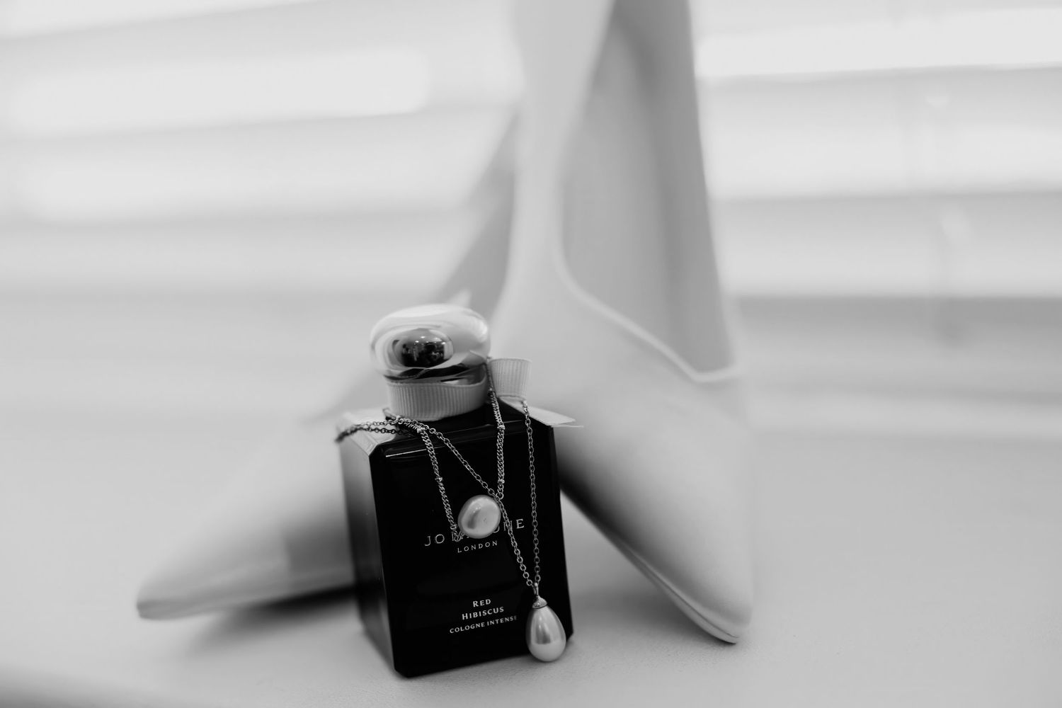 Black and white flat lay of bridal accessories including perfume bottle, pearl necklace, and white heels.