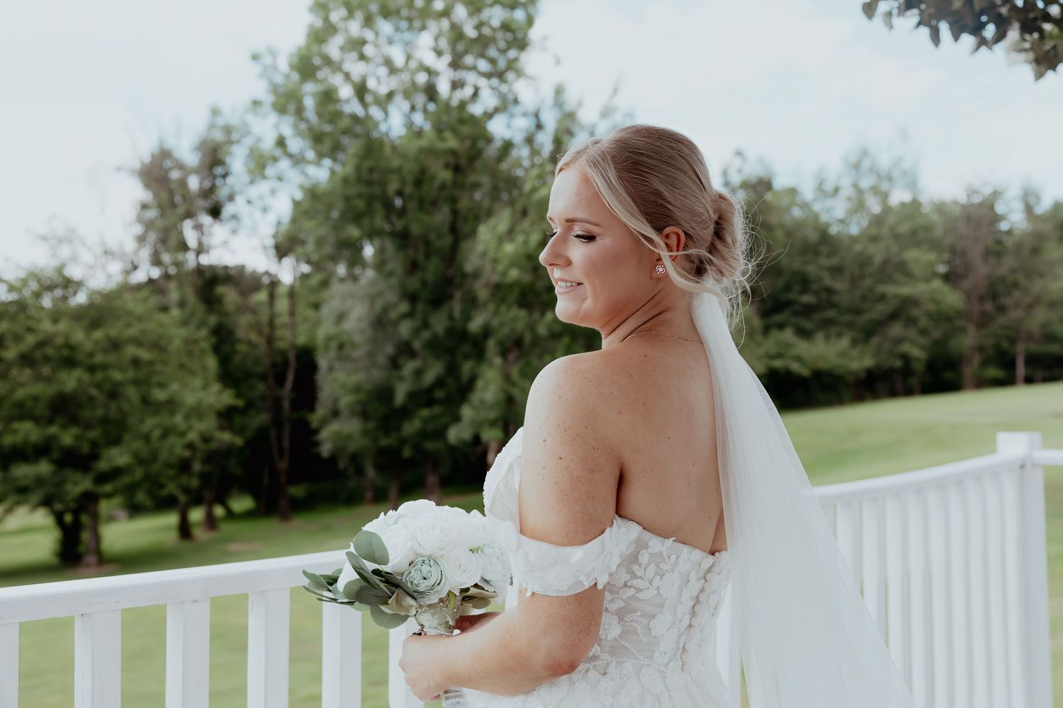 Bride in off-shoulder lace gown holding white bouquet with veil, posing on white porch railing outdoors.