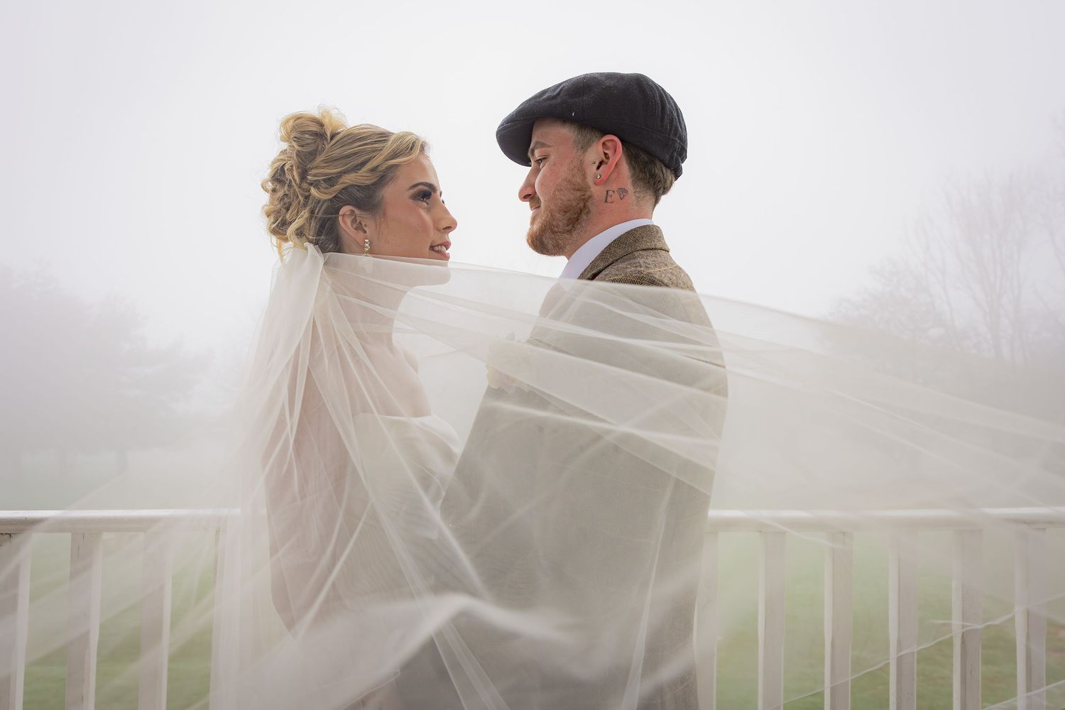 Bride and groom wrapped together in a flowing veil on a misty white balcony, gazing into each other's eyes.