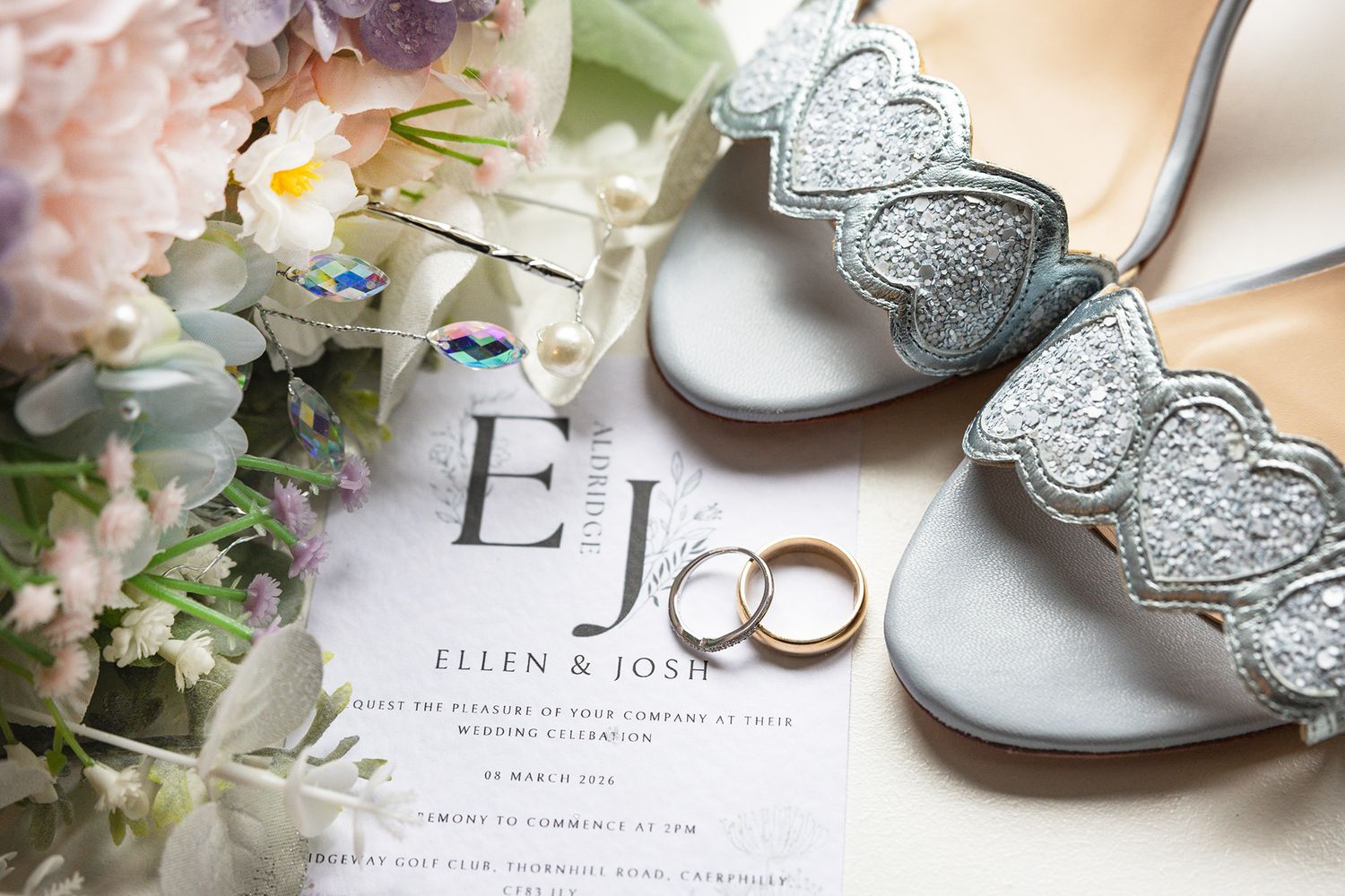 Wedding flat lay featuring invitation for Ellen and Josh, gold rings, silver heels and flowers.