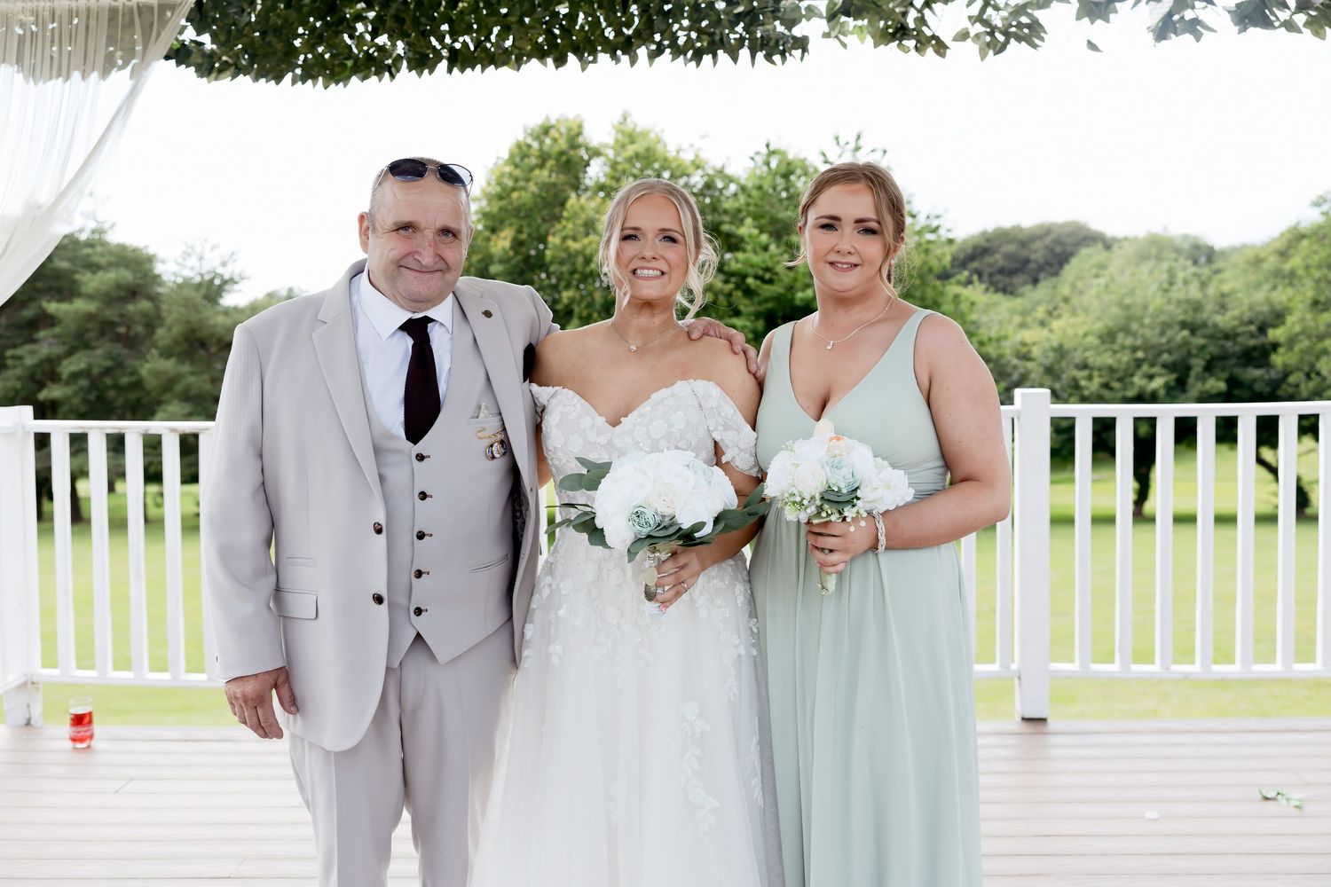 Bride in lace wedding gown poses with man in grey suit and woman in mint bridesmaid dress outdoors.