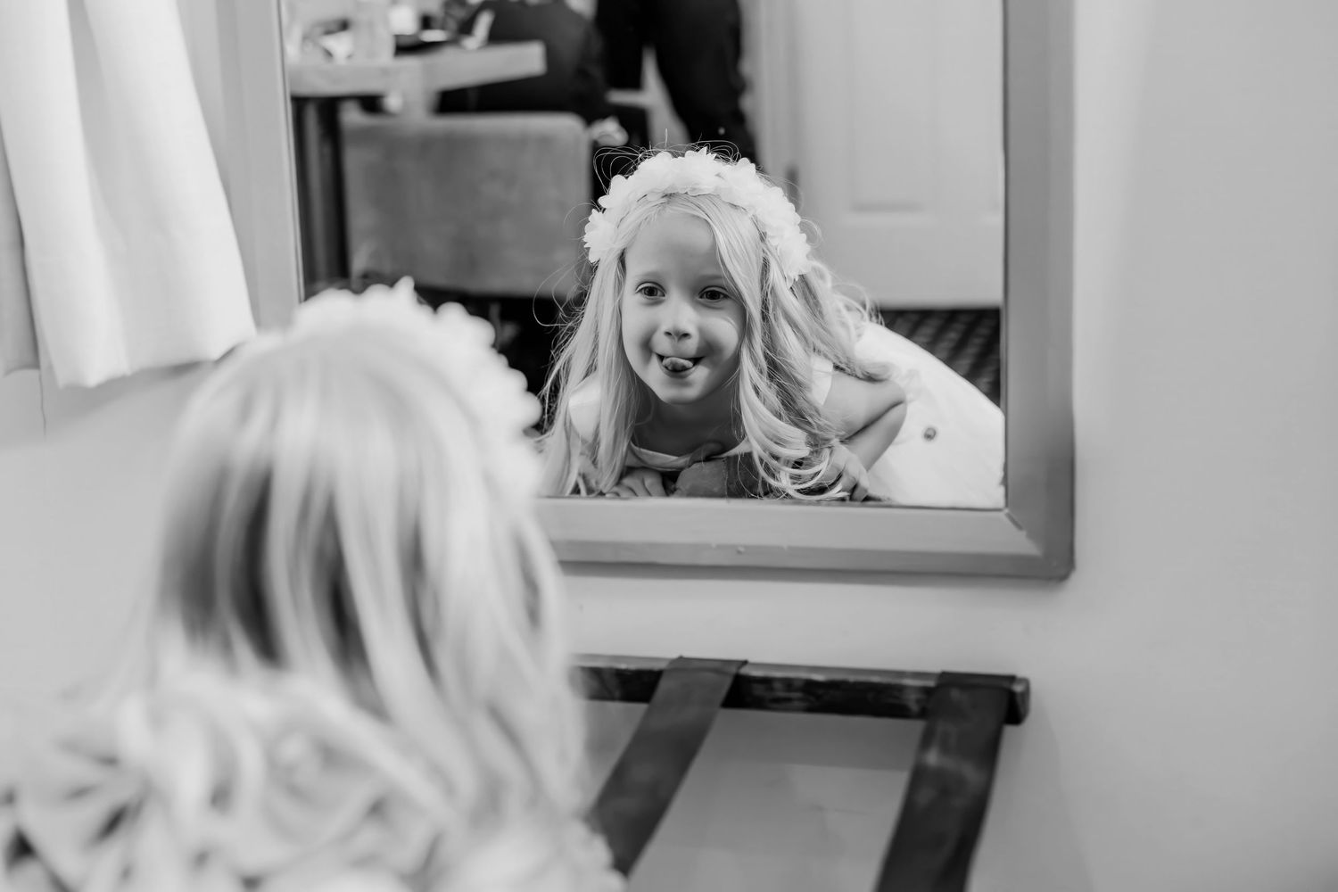 A flower girl with a floral crown smiles at her reflection in a mirror in this black and white photo.