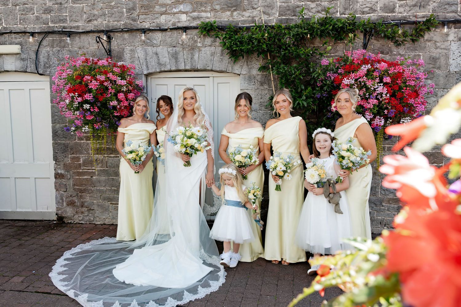 Bride in white gown with bridesmaids in yellow dresses holding bouquets, posing with flower girls outside stone venue.