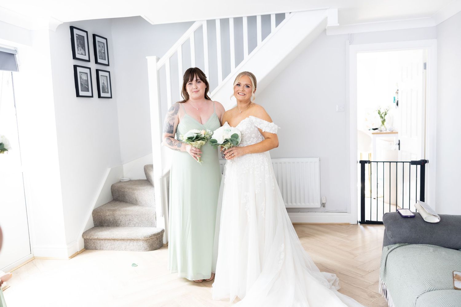 Bride in off-shoulder gown poses with bridesmaid in sage green dress by staircase.