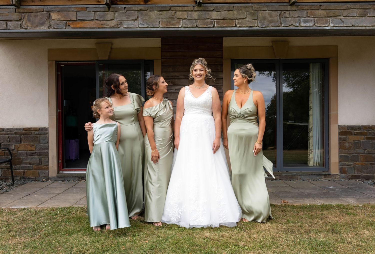 Bride in white gown poses with three bridesmaids in sage green dresses and flower girl outside stone building.