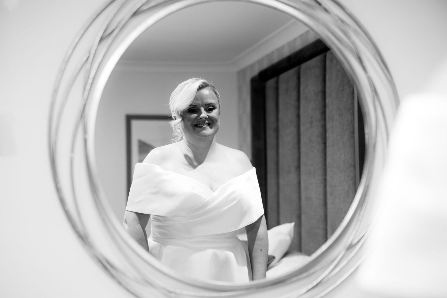 Black and white reflection of a smiling bride in a round mirror while getting ready.