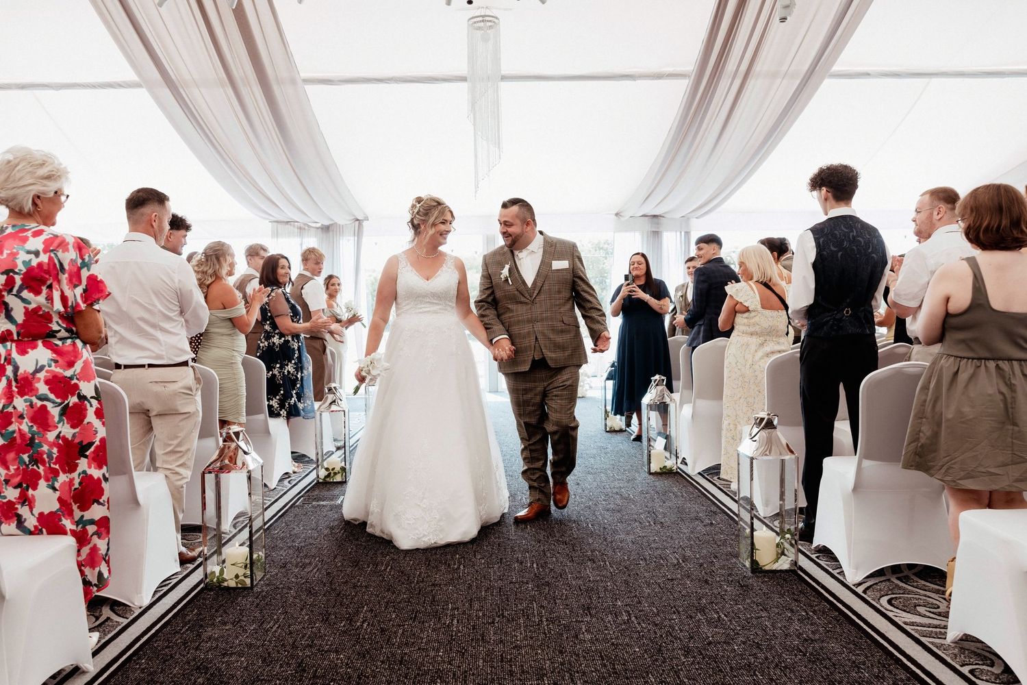Bride and groom walk down the aisle smiling at outdoor wedding ceremony with guests seated on both sides.