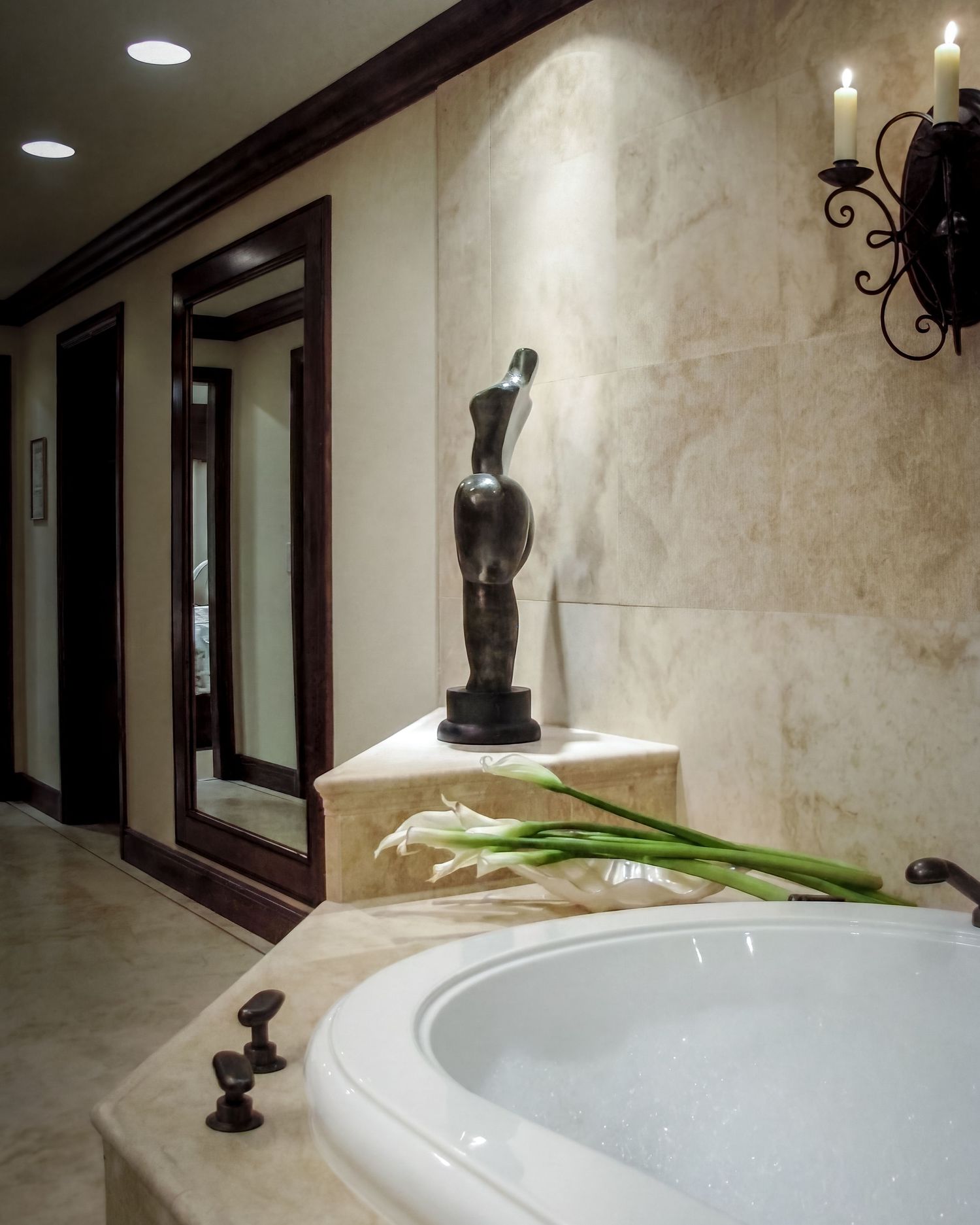 Luxurious bathroom with soaking tub, abstract sculpture, white calla lilies, marble walls, and elegant iron wall sconce.