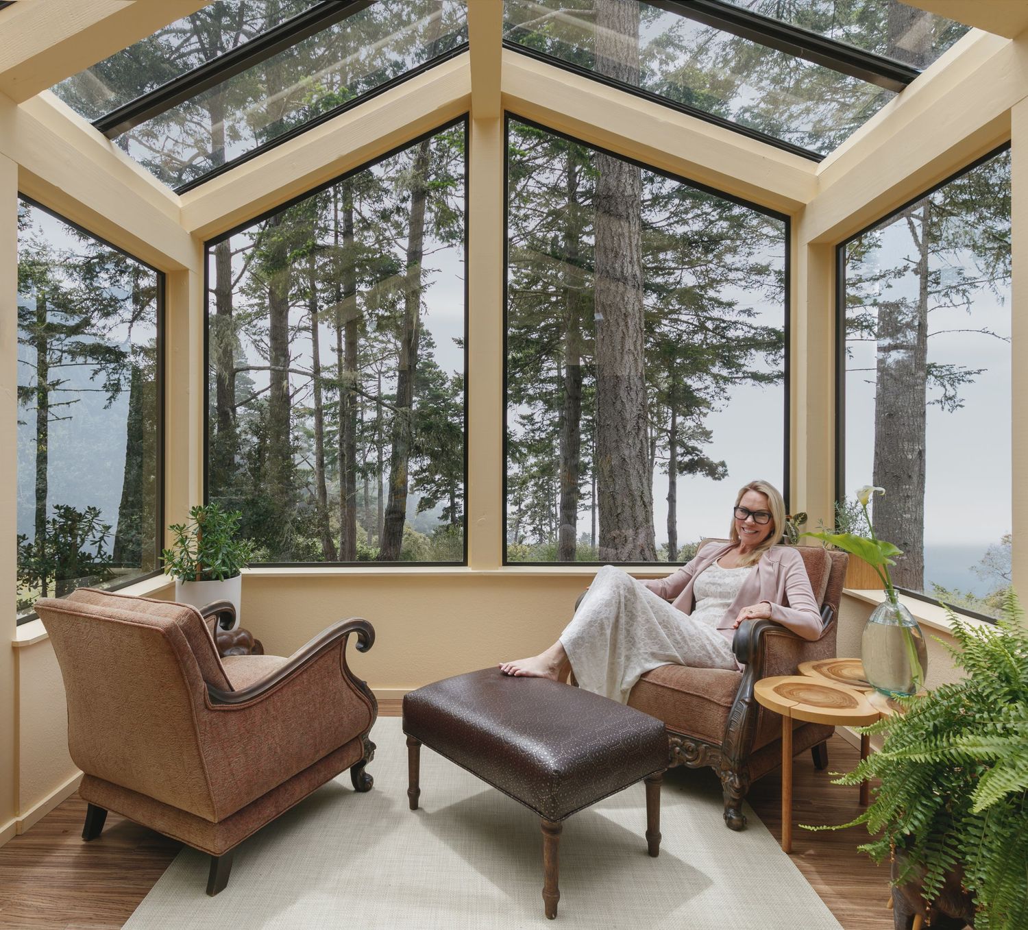 Woman relaxing in sunroom with floor-to-ceiling windows overlooking tall trees and scenic forest landscape.