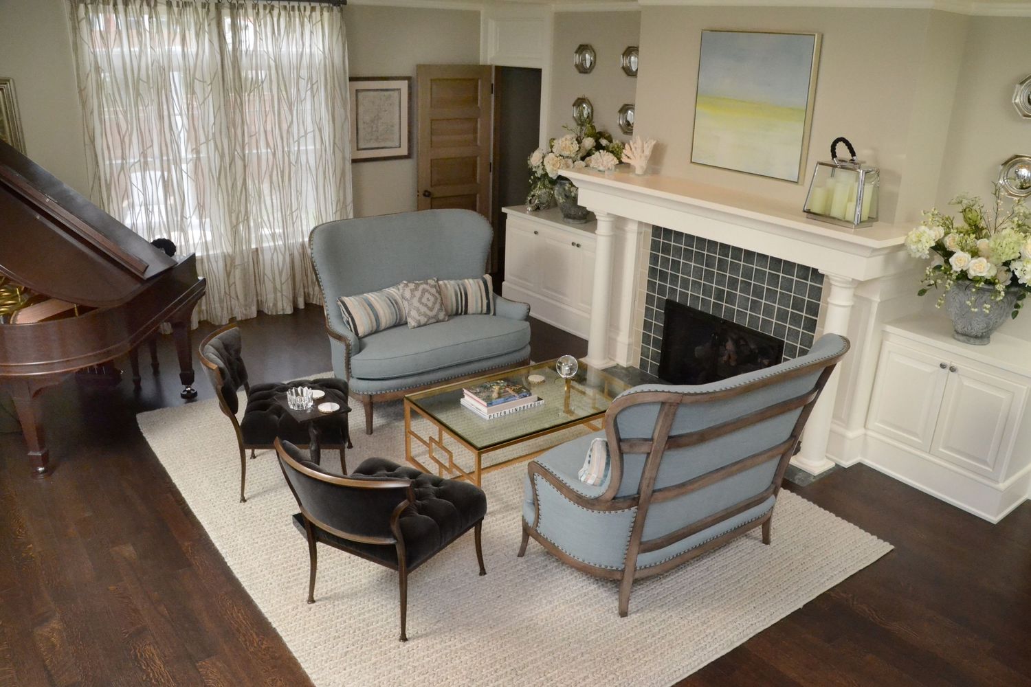 Elegant living room with blue upholstered chairs, white fireplace, glass coffee table, and grand piano on hardwood floors.