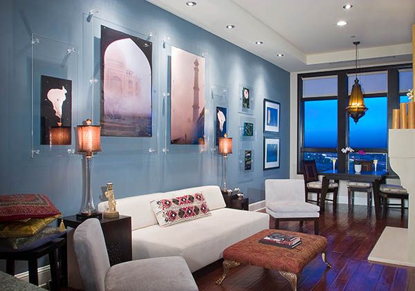 Modern living room with blue walls, white sofa, artwork gallery, dark wood floors, and elegant dining area with pendant lights.