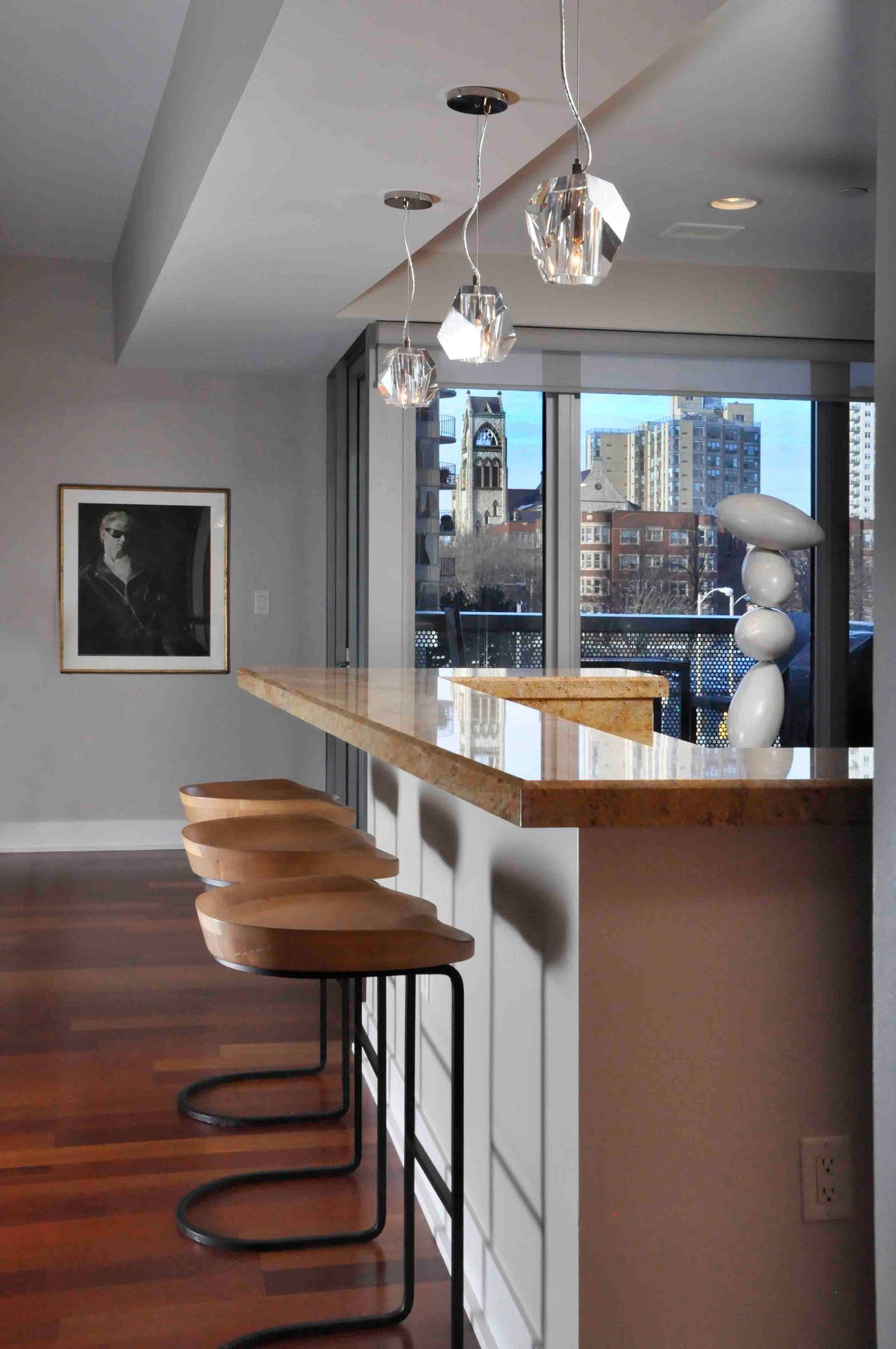 Modern kitchen bar with wood stools, pendant lights, marble countertop, and city skyline view through large windows.