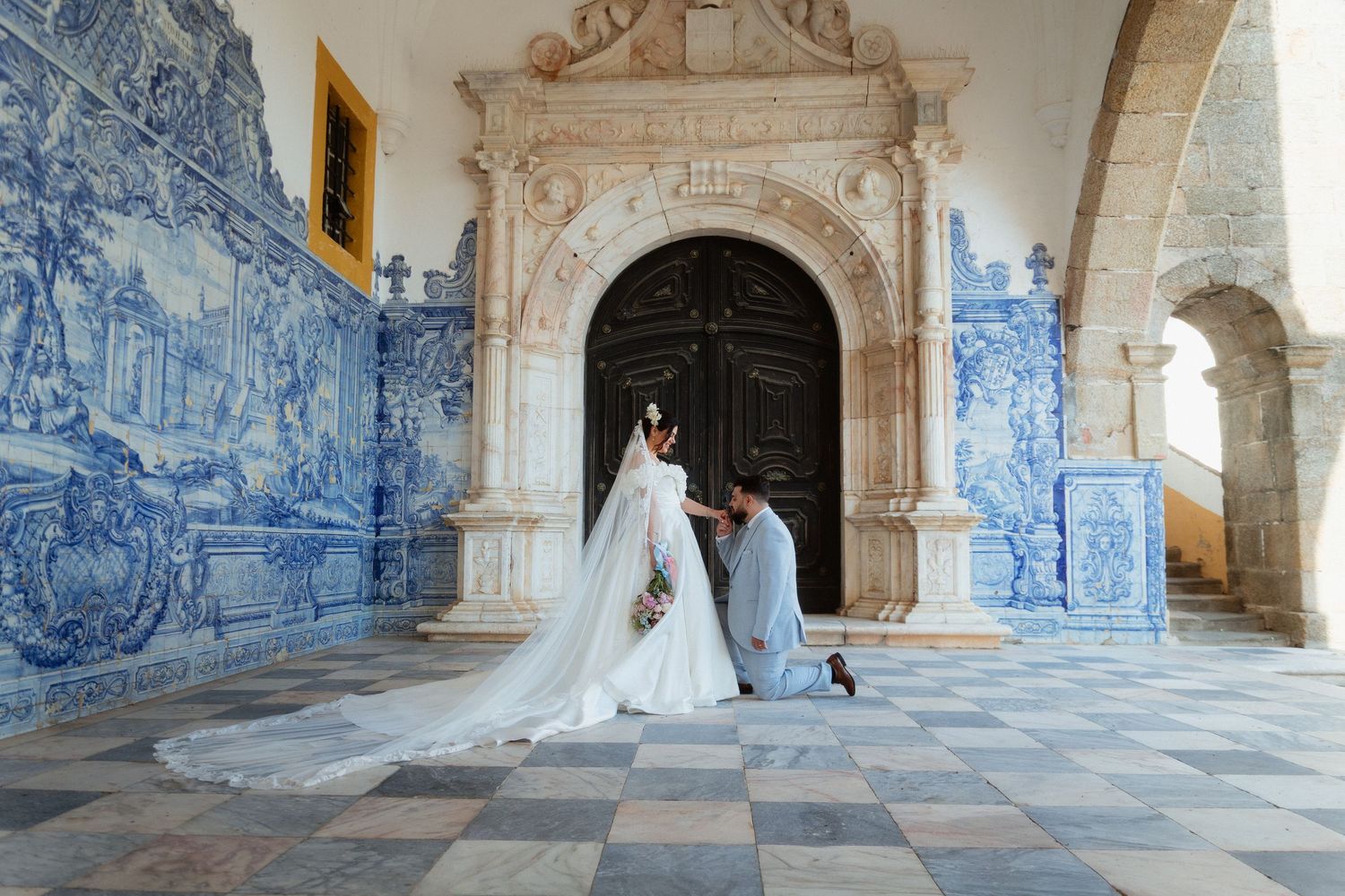 Noivos diante de uma porta e azulejos azuis e brancos, noivo ajoelhado beija mão da noiva, em Mosteiro de São Bernardo, Portalegre.