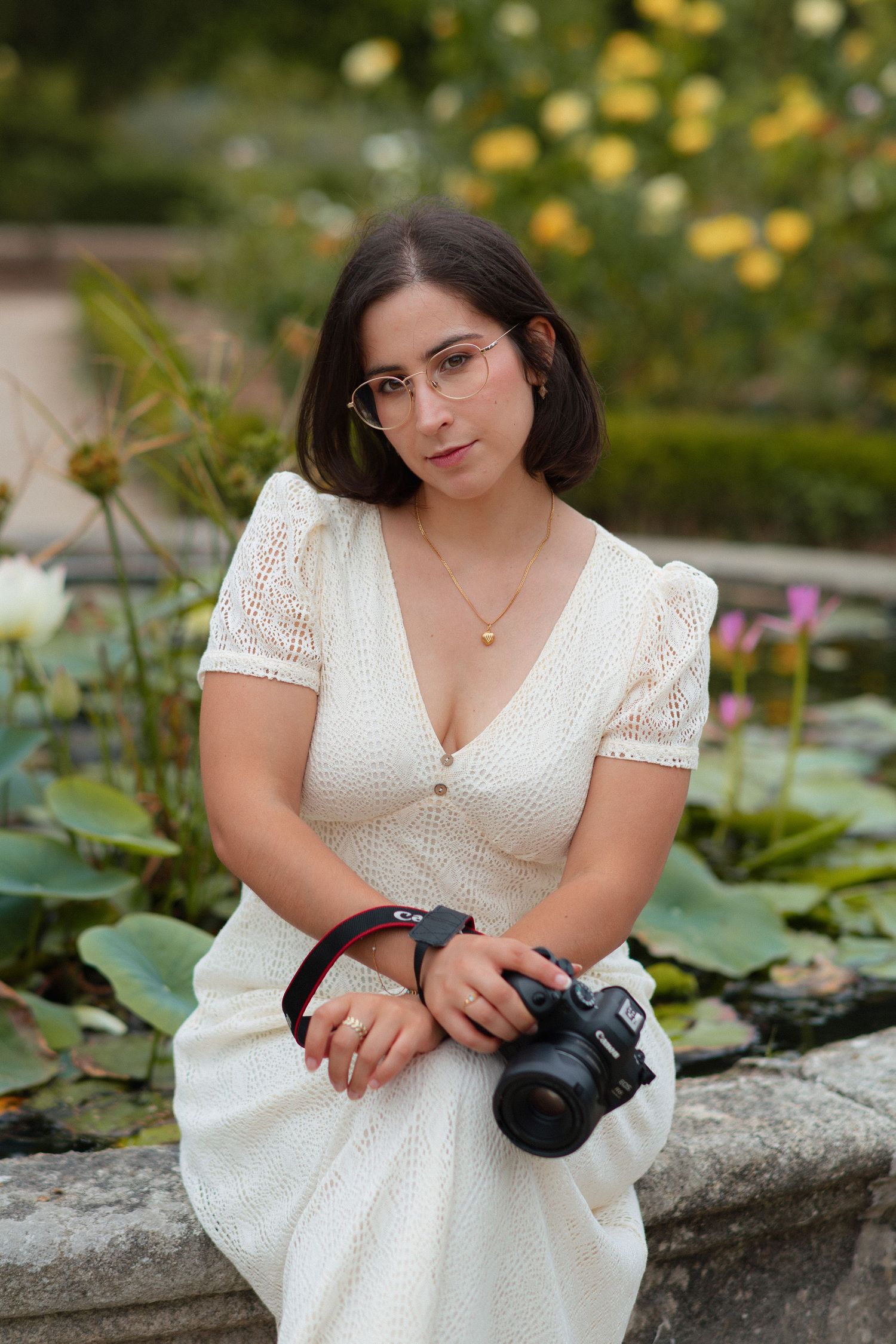Cristiana Ribeiro sentada na beira de uma fonte, com água, vegetação e flores, a segurar câmara na mão, no Jardim Botânico do Porto.