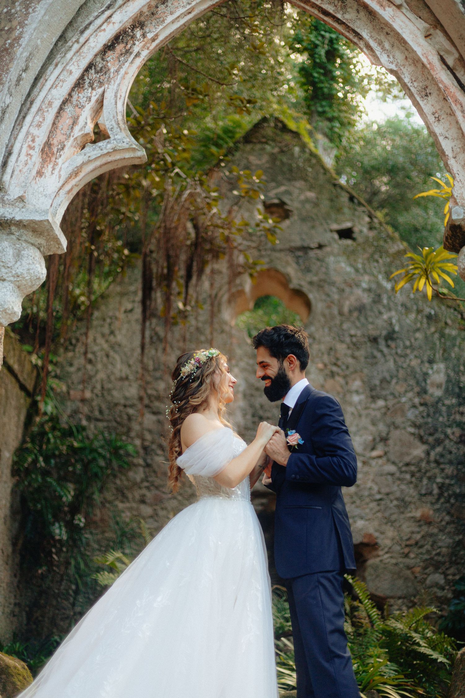 Noivos encontram-se e dão mãos sob um arco de pedra coberto de vegetação, em ruínas de capela, em Monserrate, Sintra.