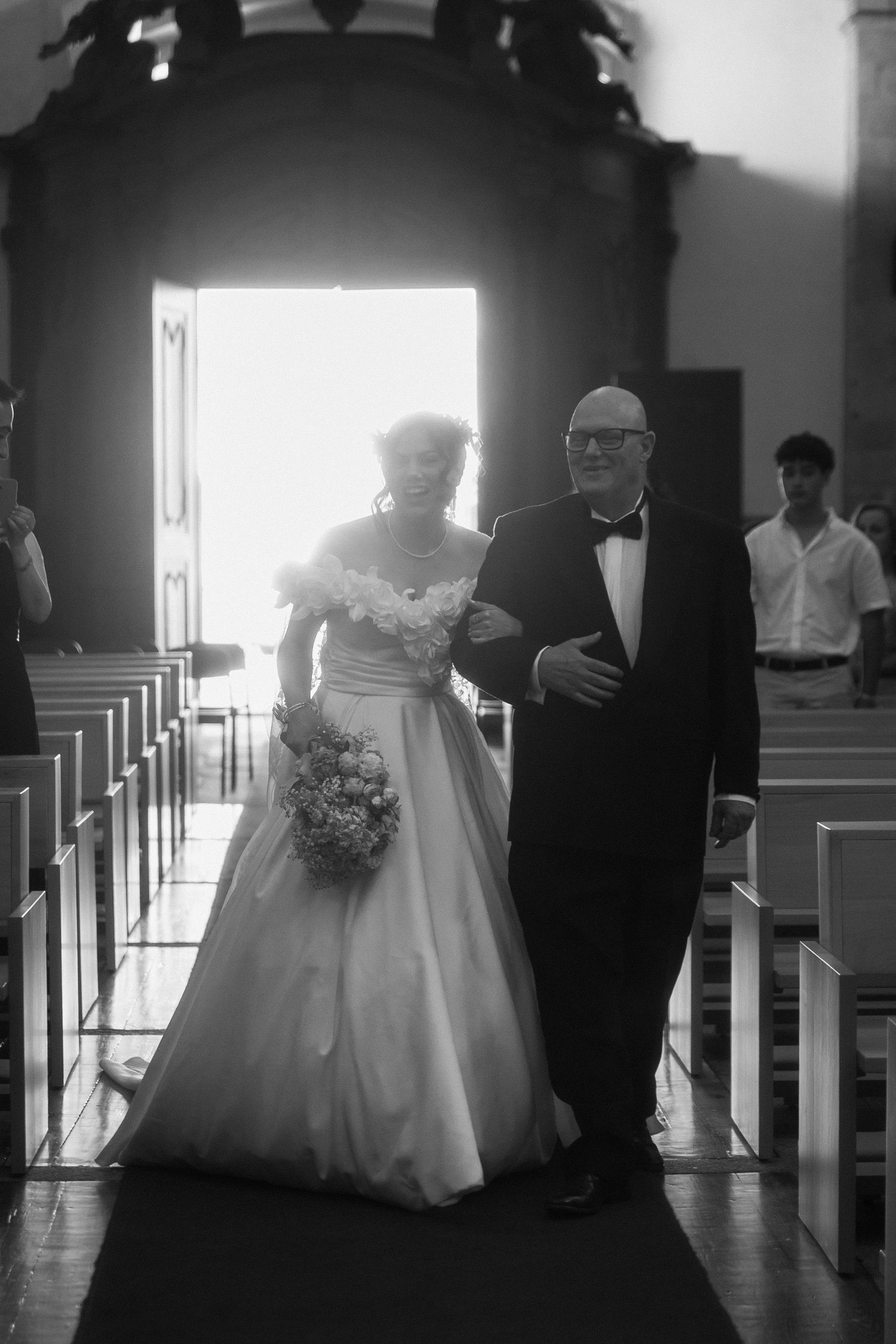 Noiva entra na igreja acompanhada pelo pai, sorrindo durante um momento emotivo antes da cerimónia de casamento.