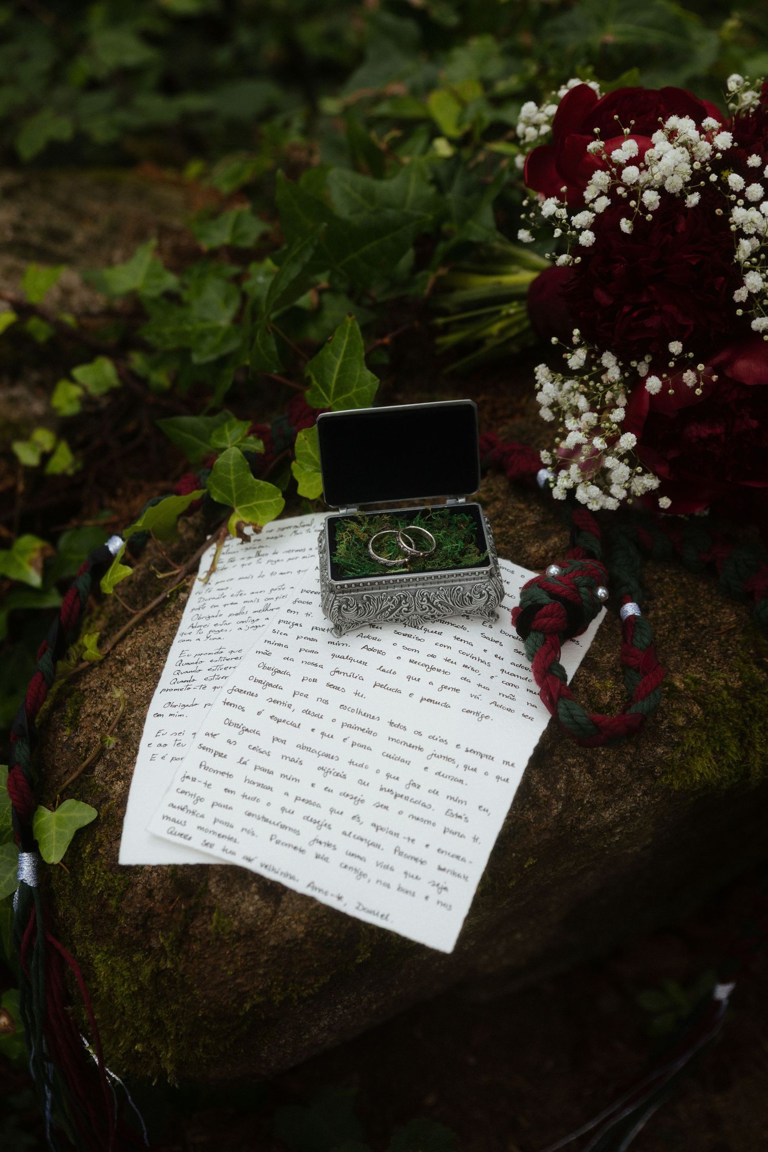 Votos escritos, anéis, bouquet e corda de handfasting dispostos sob uma pedra, rodeados por elementos naturais, numa floresta em Sintra.
