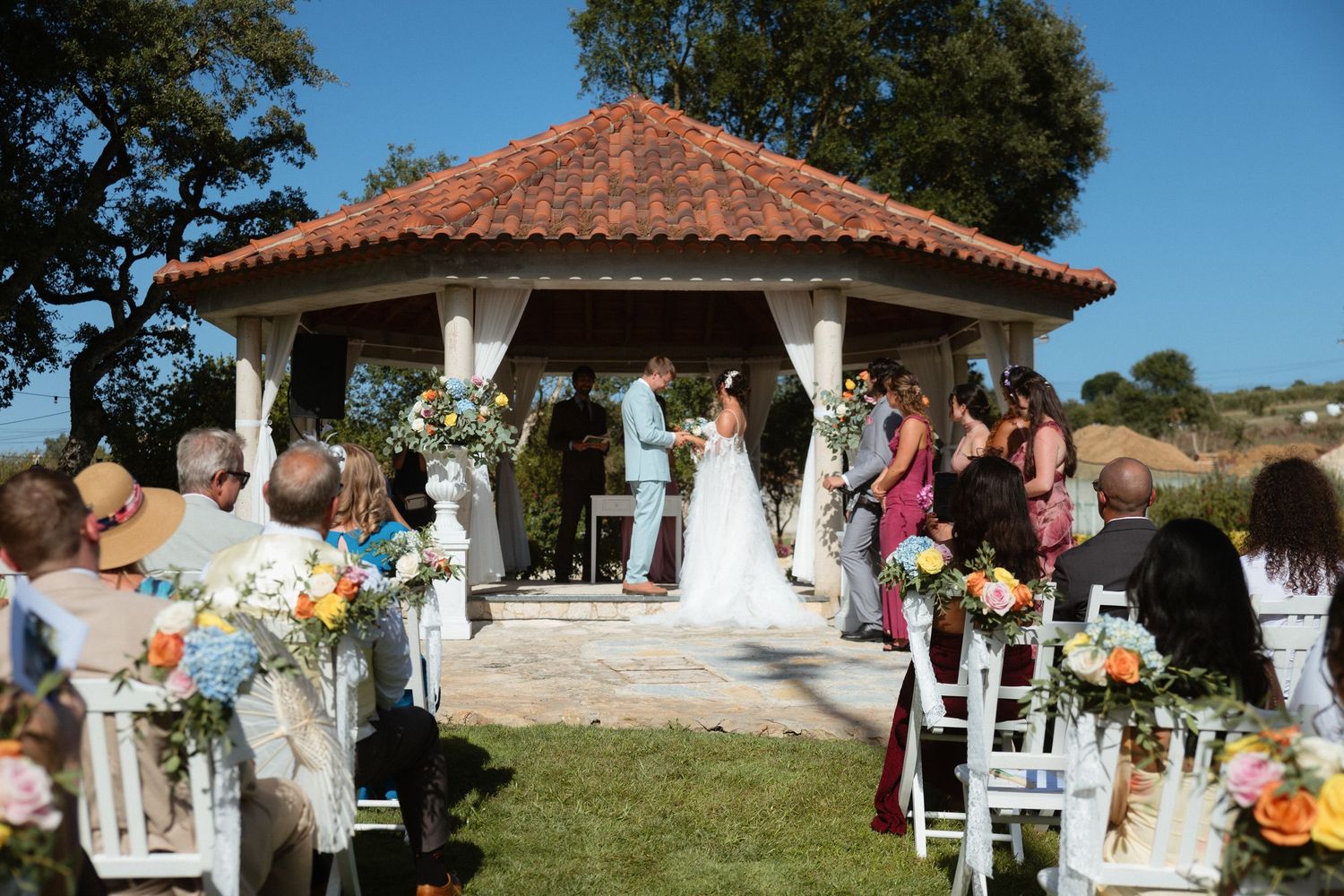Casamento em jardim, com noivos junto ao altar, convidados em cadeiras brancas, rodeados de flores coloridas, em Alcobaça.