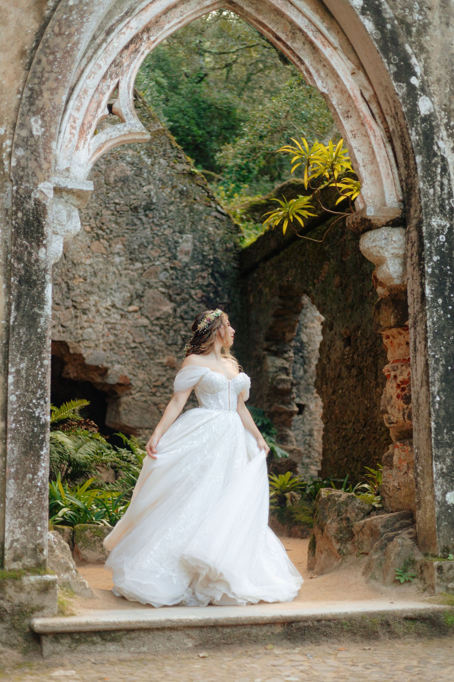 Noiva de vestido posa sob um antigo arco de pedra coberto de vegetação, num cenário romântico de ruínas de capela, em Monserrate, Sintra.