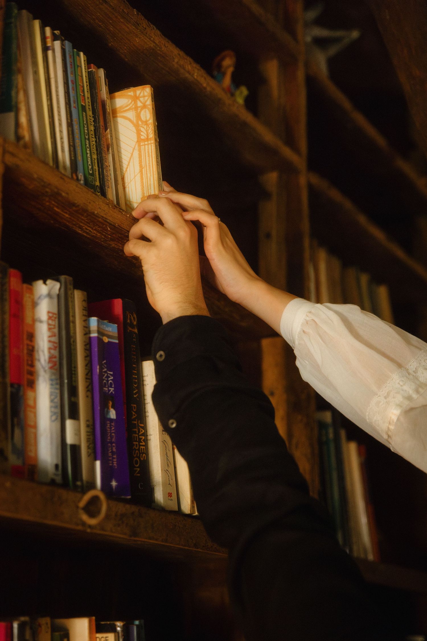 Mãos de noivos pegam num livro antigo numa estante num biblioteca antiga, num detalhe inspirado em histórias e objetos com memória.