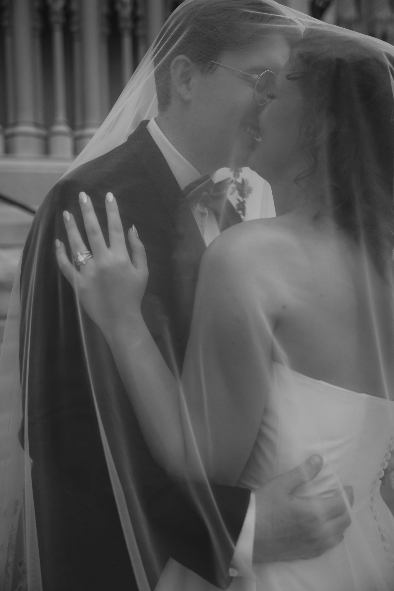 Black and white wedding photo of bride and groom kissing under veil, bride wearing diamond ring.