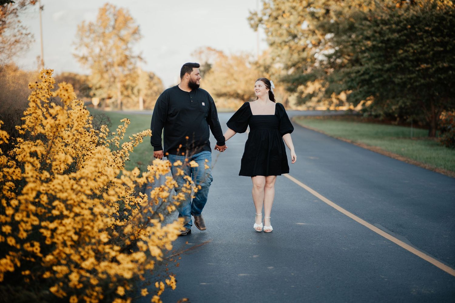 A couple walks together on a paved path bordered by vibrant yellow wildflowers on an autumn afternoon.